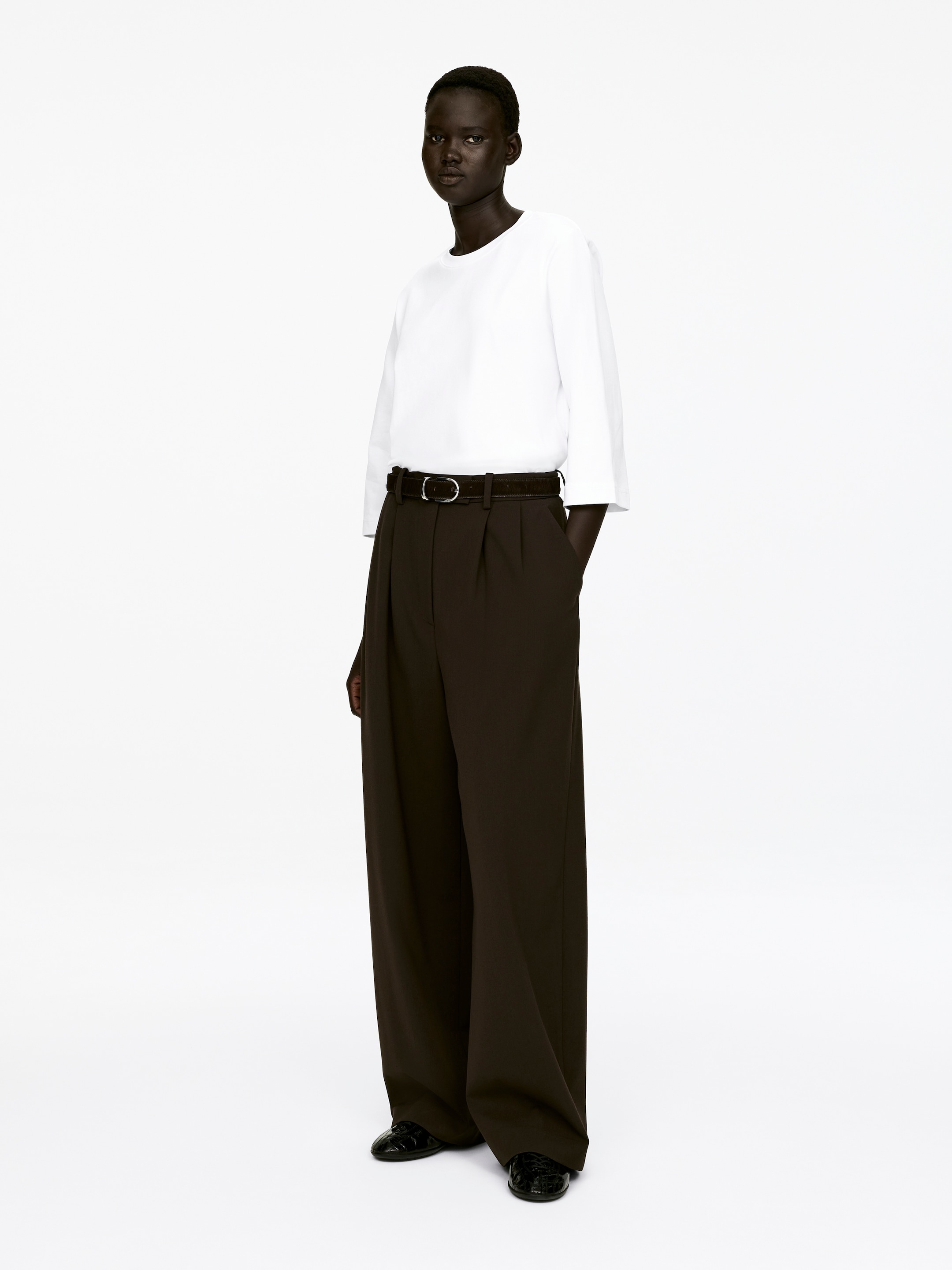 Agrandir l'image: Un T-shirt blanc à col rond, confectionné en jersey de coton épais et frais, est porté par le mannequin. Il présente des manches trois-quarts et est rentré dans un pantalon large et sombre, maintenu par une ceinture.