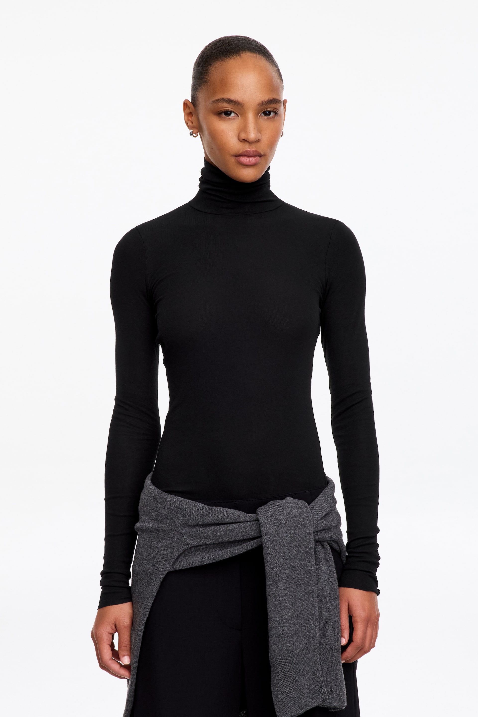 Größeres Bild anzeigen: Das Model trägt ein schwarzes Jersey-Oberteil mit langen Ärmeln und Rollkragen. Das tailliert geschnittene Oberteil ist mit einer schwarzen Hose und einem um die Taille gebundenen grauen Pullover kombiniert.