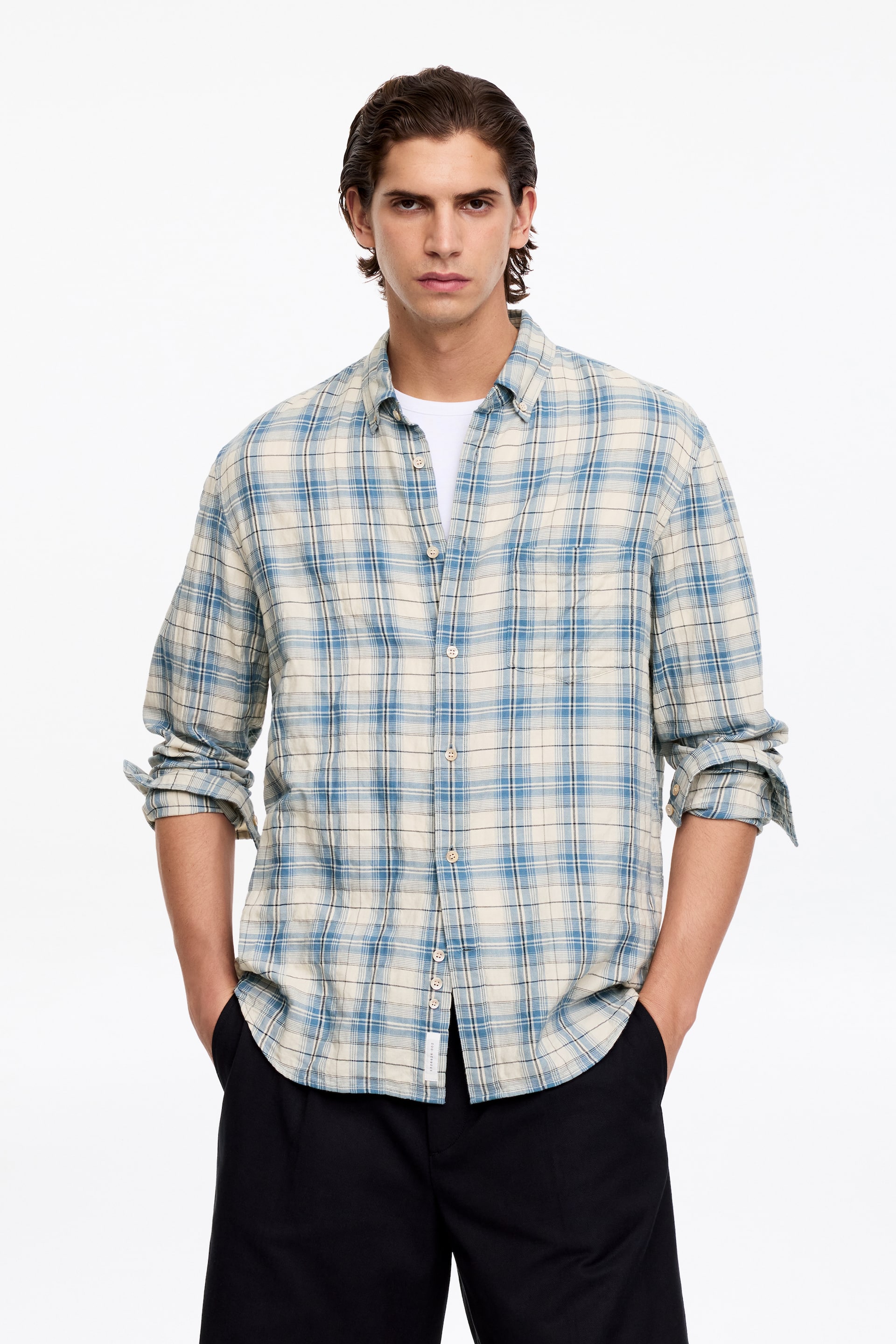 Visa större bild: En blå och beige rutig skjorta är vänd framåt, med en button-down krage och knäppning framtill. Dess långa ärmar är uppkavlade till underarmarna, och den bärs öppen över en vit t-shirt, vilket ger en avslappnad passform.