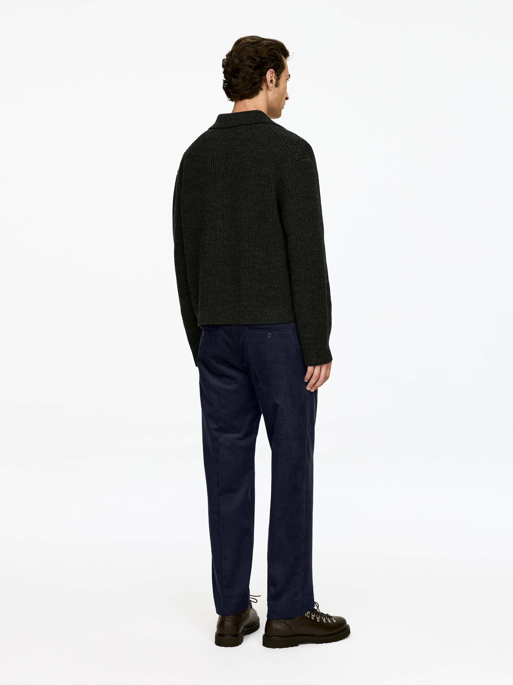 Ingrandisci l'immagine: Un maglione polo a coste di colore verde scuro viene presentato da una prospettiva posteriore leggermente angolata, caratterizzato da un colletto polo e una texture a coste verticali. È abbinato a pantaloni di velluto a coste blu navy e stivali con lacci marrone scuro.