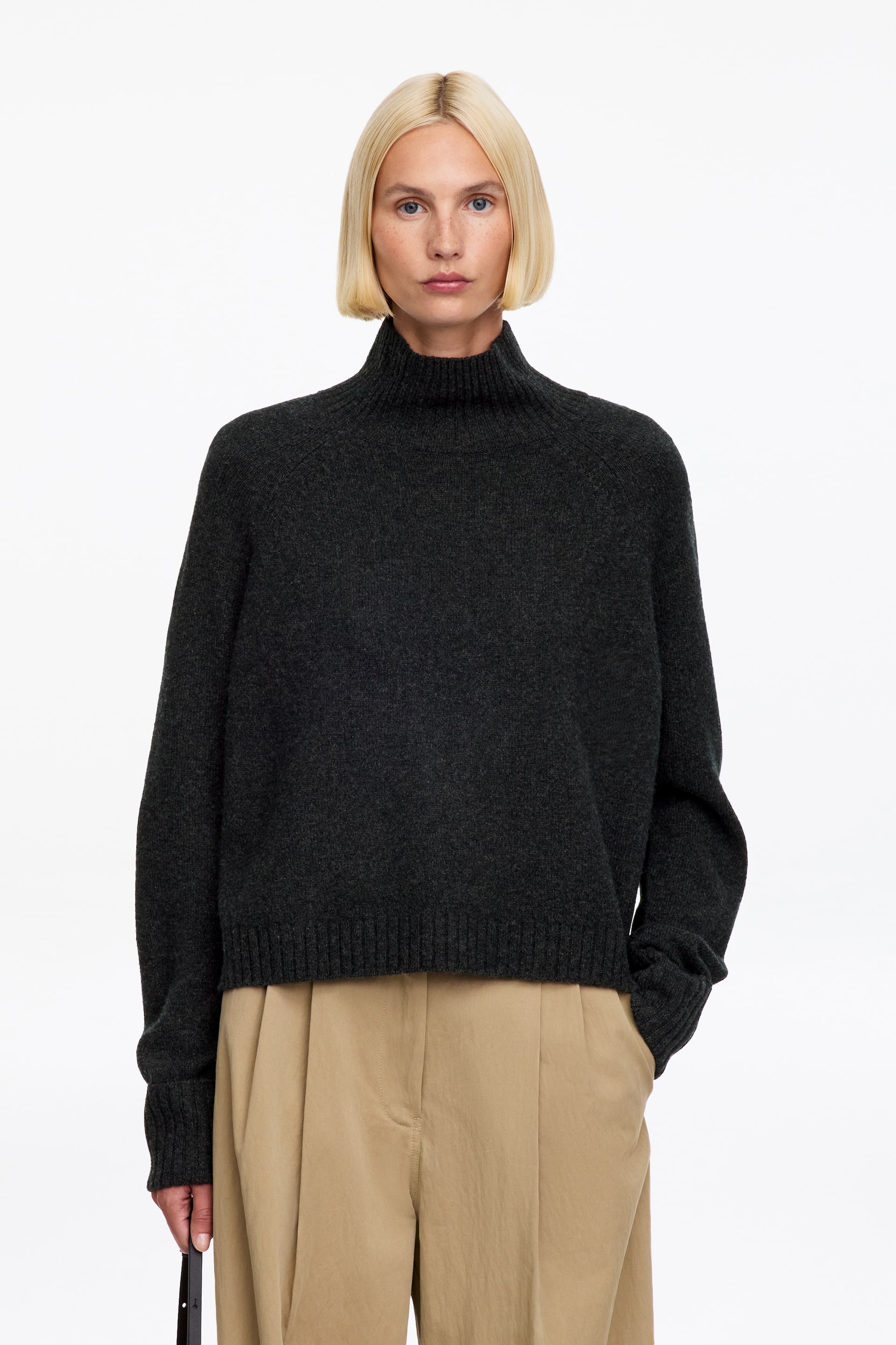 Vezi imaginea mai mare: Un pulover cu guler înalt tricotat, de culoare gri închis, este purtat de model și prezintă un decolteu înalt cu model rib, mâneci raglan și margine ribată. Puloverul este stilizat cu pantaloni bej, plisați, iar modelul își ține o mână în buzunarul pantalonilor.