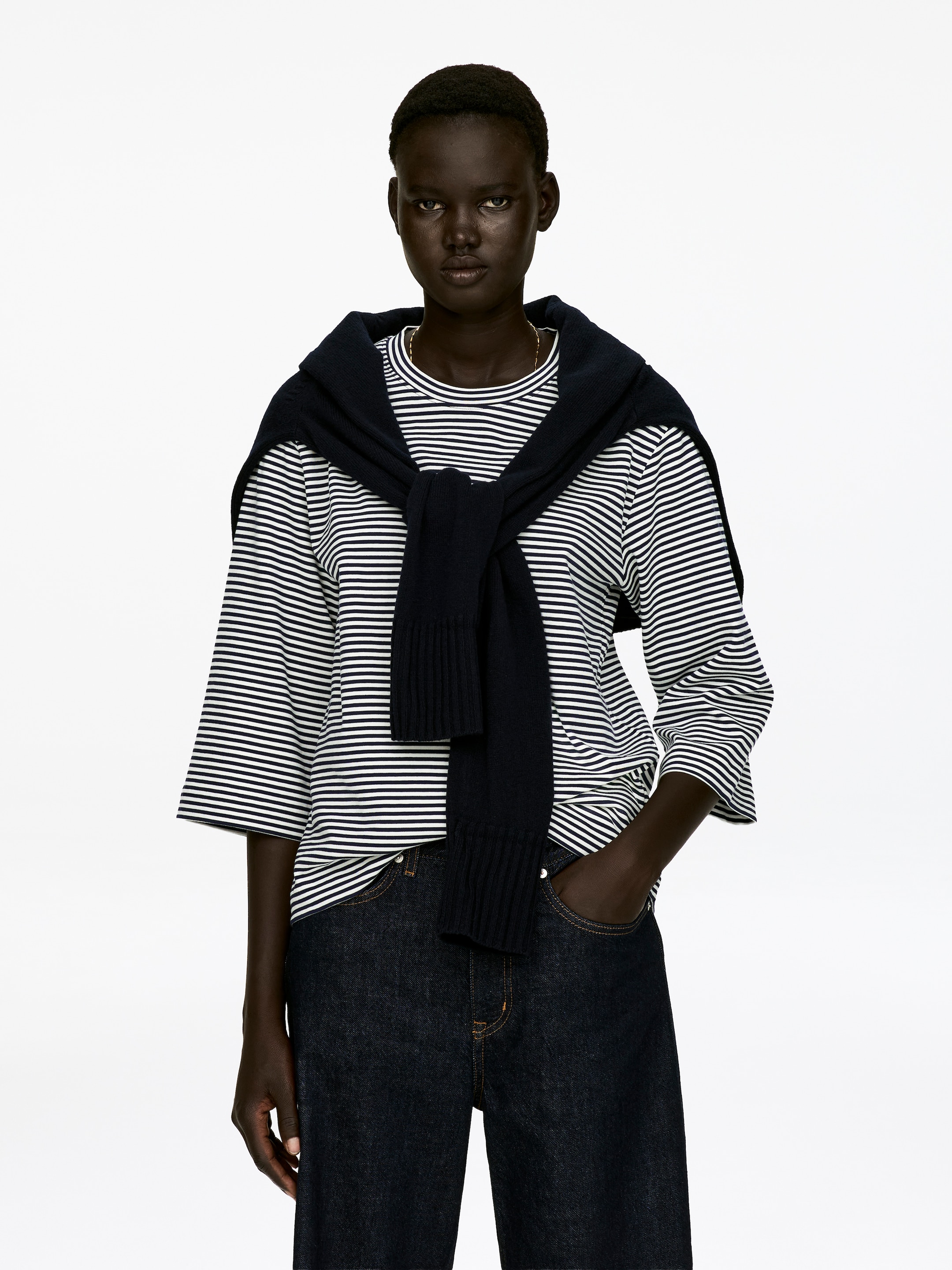 Agrandir l'image: Un T-shirt à col rond confectionné dans un jersey de coton épais et net est porté par le mannequin. Il est stylisé avec des manches 3/4 et arbore des rayures horizontales noires et blanches, avec un pull bleu marine noué autour des épaules du mannequin, le superposant.