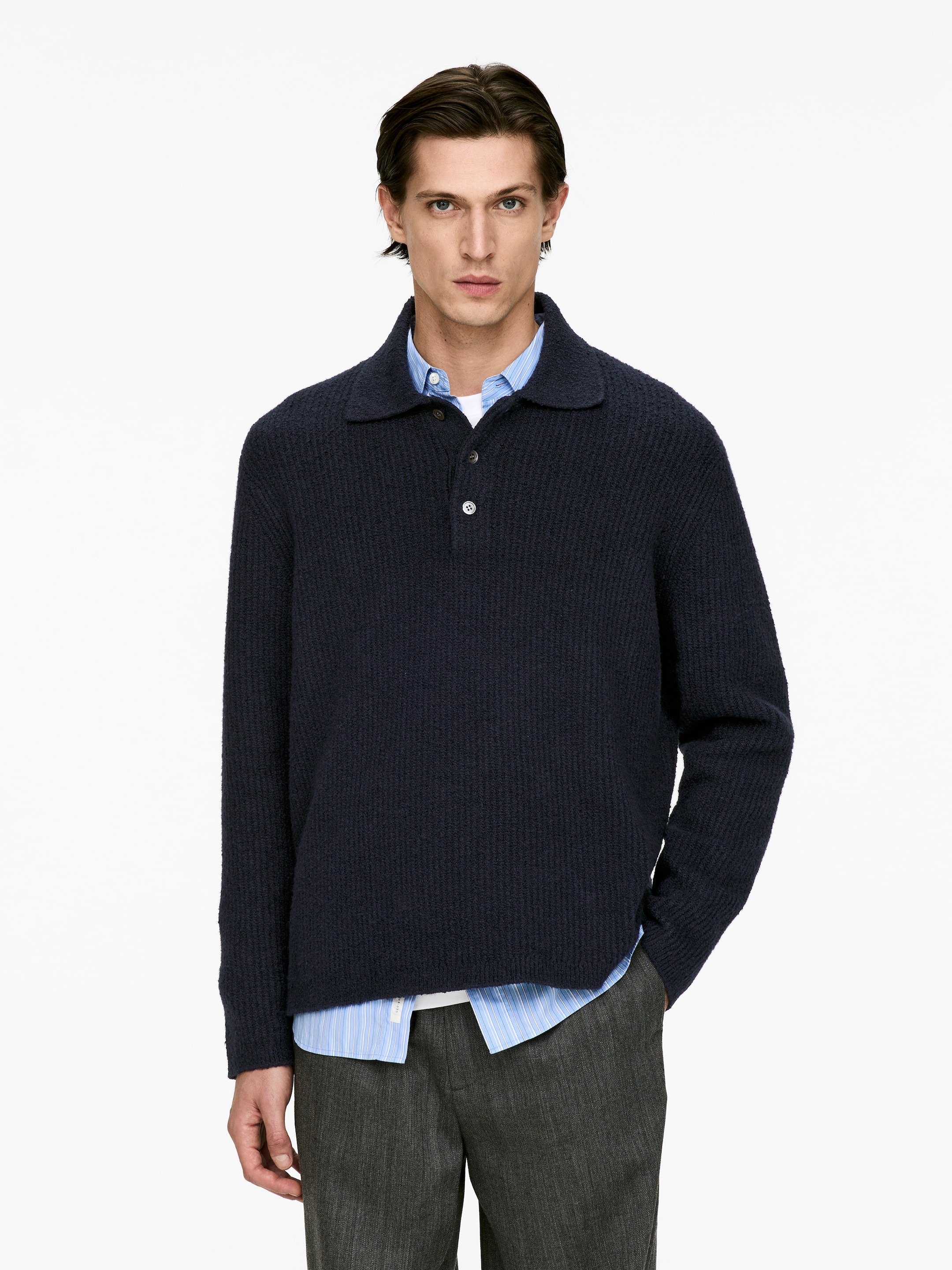 Vis større bilde: En marineblå teksturstrikket polosweater vender fremover, med lange ermer, en polokrage og en knappestolpe med tre lyse knapper. Den er lagd på en lyseblå og hvitstripet skjorte med krage, som er synlig ved halsen og nederkanten.