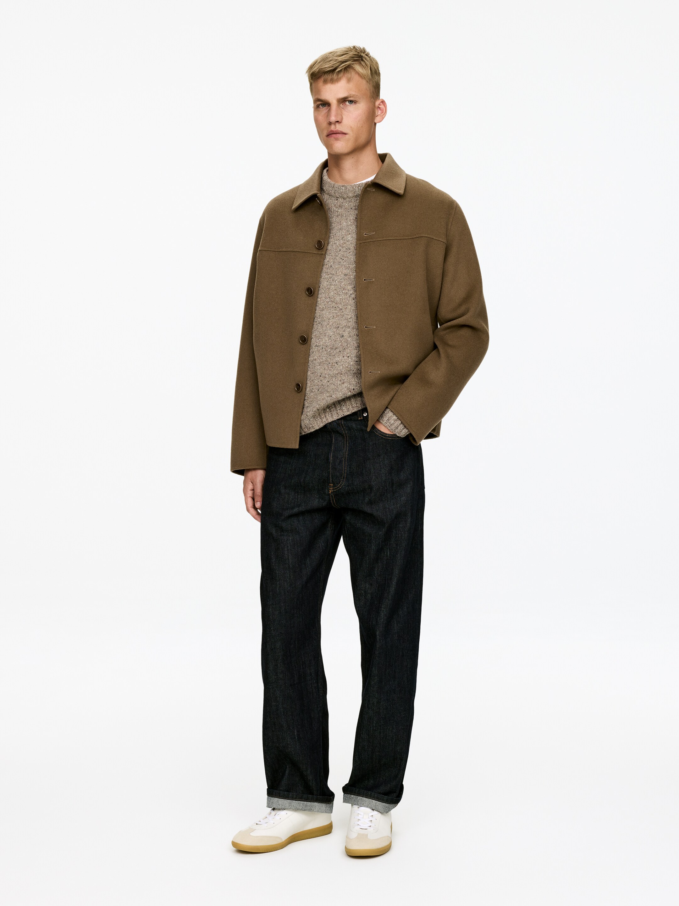 Se større billede: Et outfit består af en kort, brun jakke med krave og knaplukninger, lagt over en lysebrun sweater med crew-neck og spættet mønster. Det er parret med mørkvaskede jeans med lige ben, opsmøget ved anklerne, og hvide sneakers med gummisåler, præsenteret fra en let venstre vinkel for at fremhæve pasform og design.