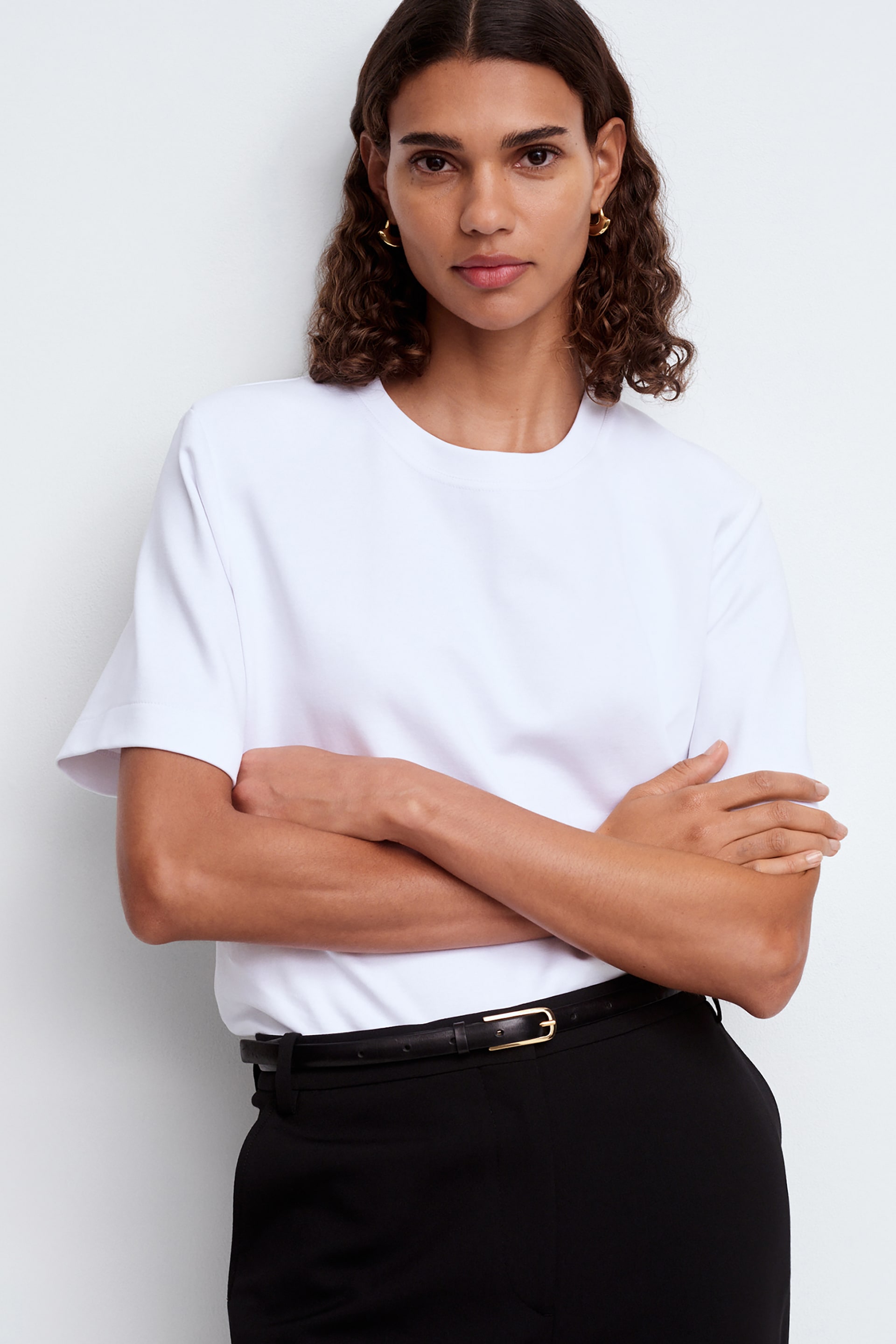 Visualizza immagine più grande: Una t-shirt bianca a girocollo è presentata frontalmente, mostrando una vestibilità rilassata e maniche corte. Questa t-shirt è infilata in pantaloni neri su misura, fissati da una cintura sottile nera con fibbia rettangolare dorata.