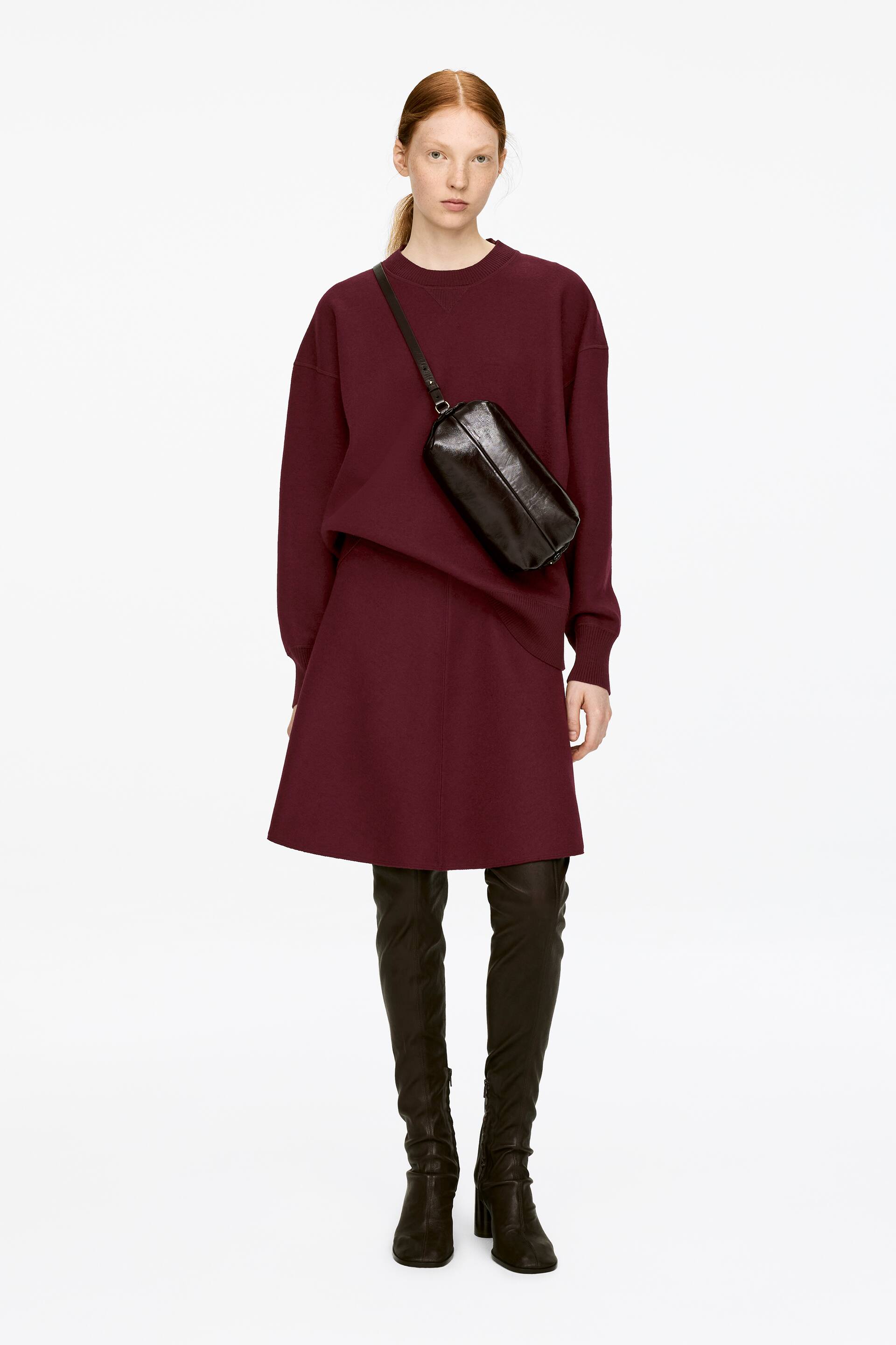 Vis større bilde: En mørkerød miniskjørt med dra-på-stil, et glatt midjebånd og en utsvingt silhuett bæres av modellen. Skjørtet er stylet med en matchende overdimensjonert genser og sorte over-kneet-støvler.