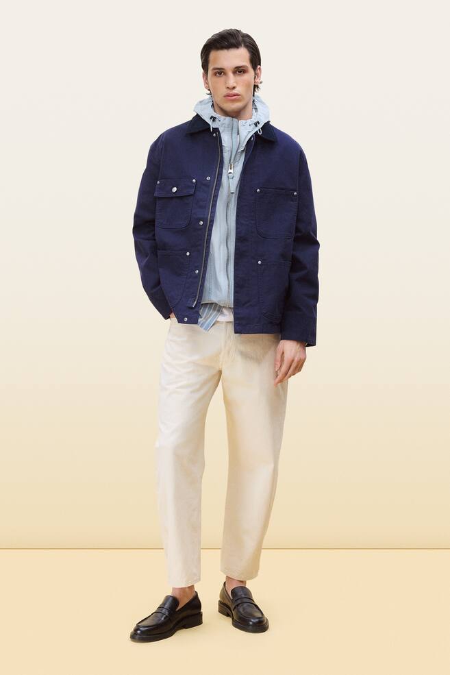 An outfit is being showcased, featuring a navy blue chore jacket with a dark contrasting collar, two chest flap pockets, and two lower patch pockets with silver-toned rivets, layered over a light blue zip-up hoodie and a light blue striped shirt. This ensemble is paired with light cream-coloured trousers and black leather loafers, and is facing forward. - 1