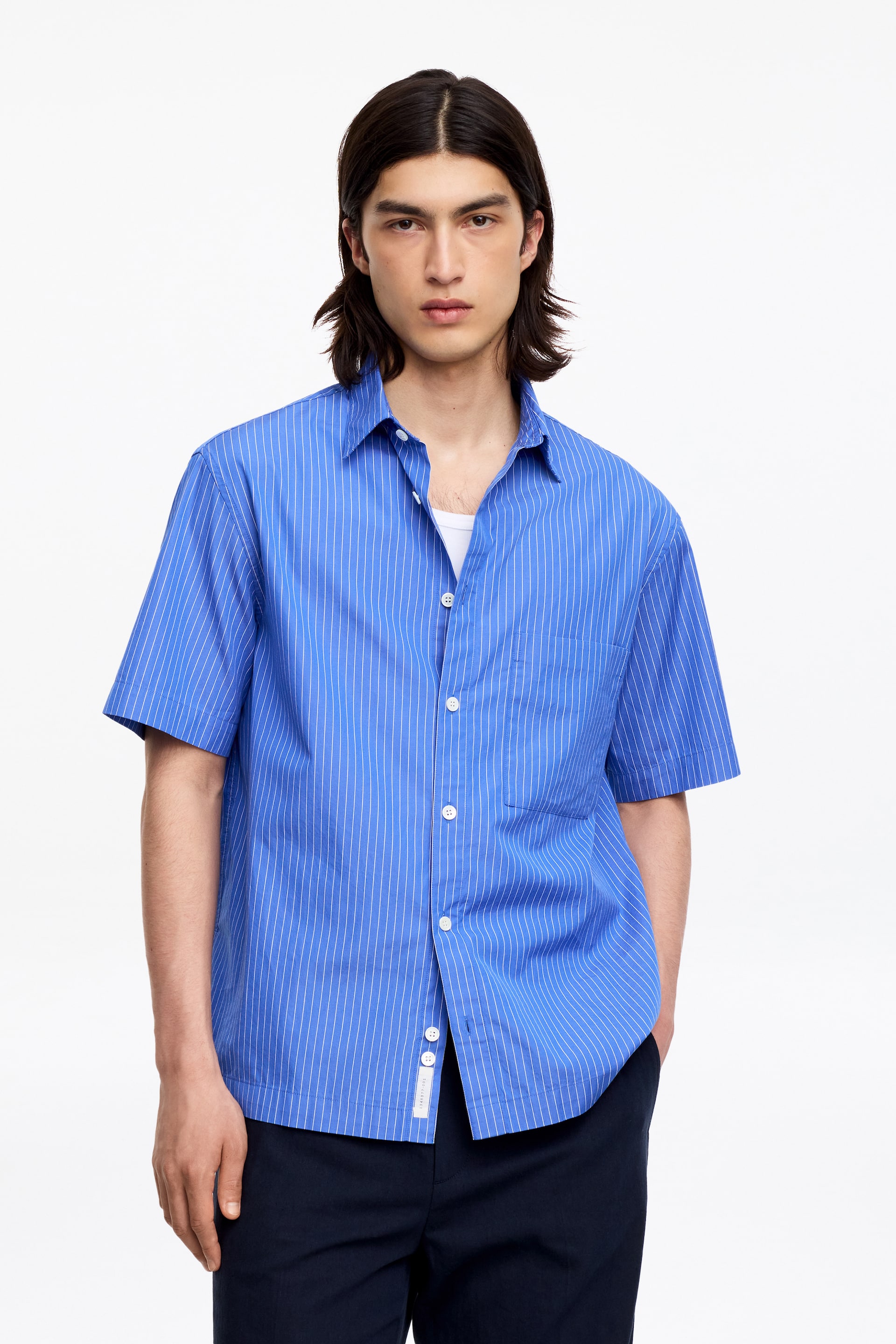 Visualizza immagine più grande: Una camicia a maniche corte blu con righe verticali bianche, caratterizzata da un colletto classico, una vestibilità rilassata e un orlo dritto, è indossata dal modello. La camicia presenta una chiusura frontale con bottoni bianchi e una tasca singola sul petto, ed è indossata parzialmente sbottonata sopra una maglietta bianca.