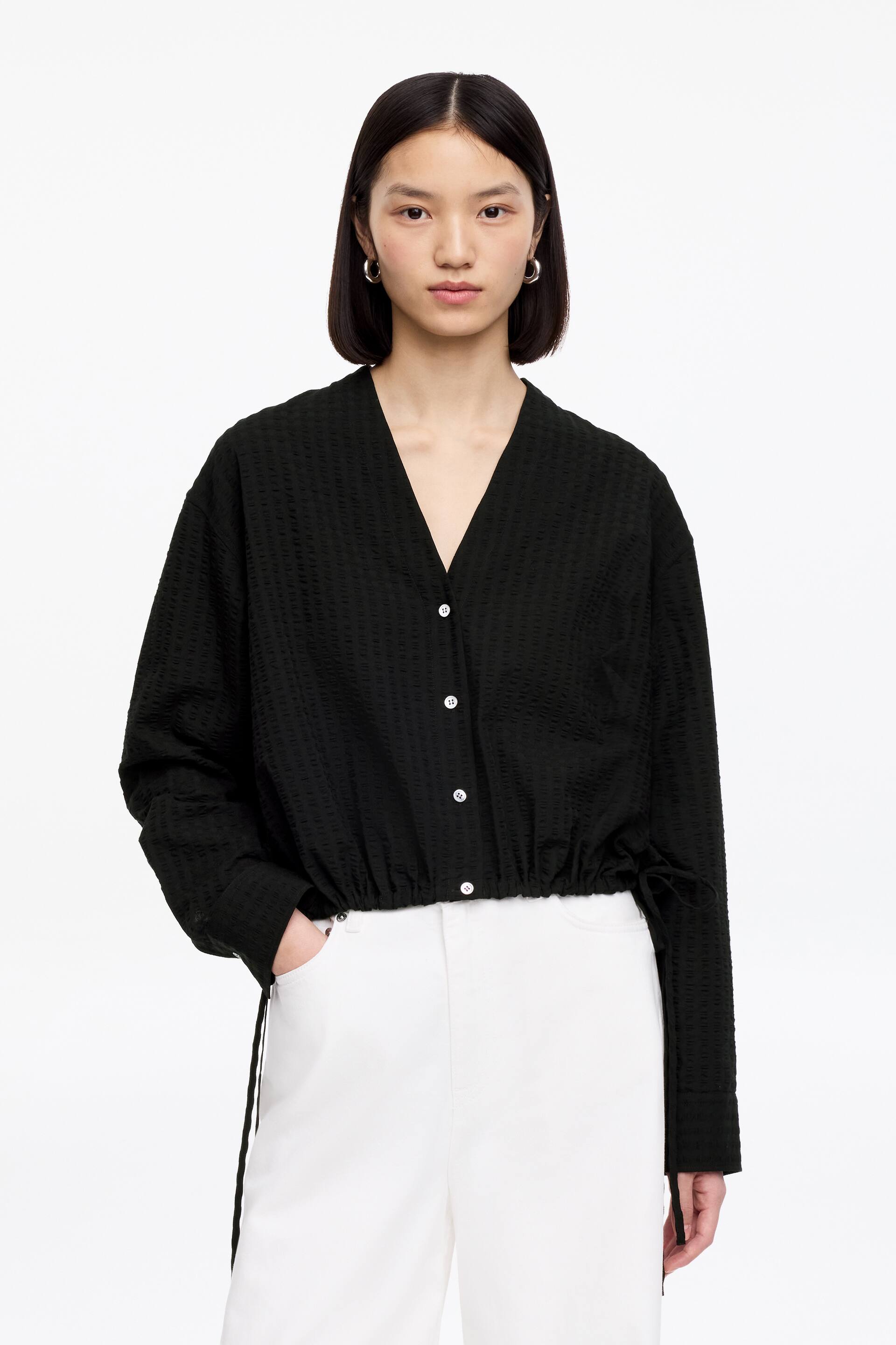 Visa större bild: En svart topp med subtil geometrisk textur, V-ringning och vit knäppning framtill. Den har långa ärmar och en åtdragen dragsko i fållen med synliga band och bärs över vita byxor.