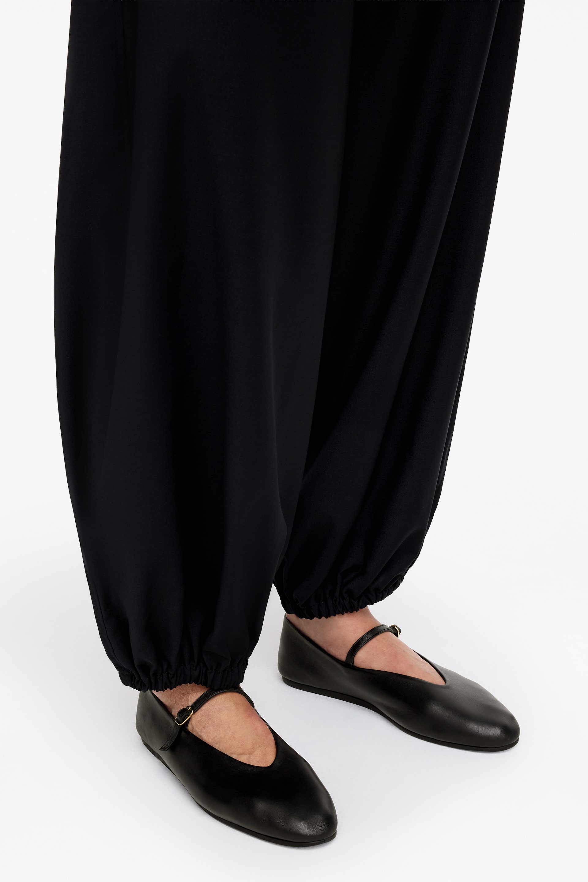 Ingrandisci l'immagine: I pantaloni neri larghi scendono fluidi verso il basso e si raccolgono alle caviglie con polsini elasticizzati. Le scarpe basse Mary Jane in pelle nera sono indossate con i pantaloni, caratterizzate da una punta affusolata e un singolo cinturino con fibbia sopra il collo del piede.