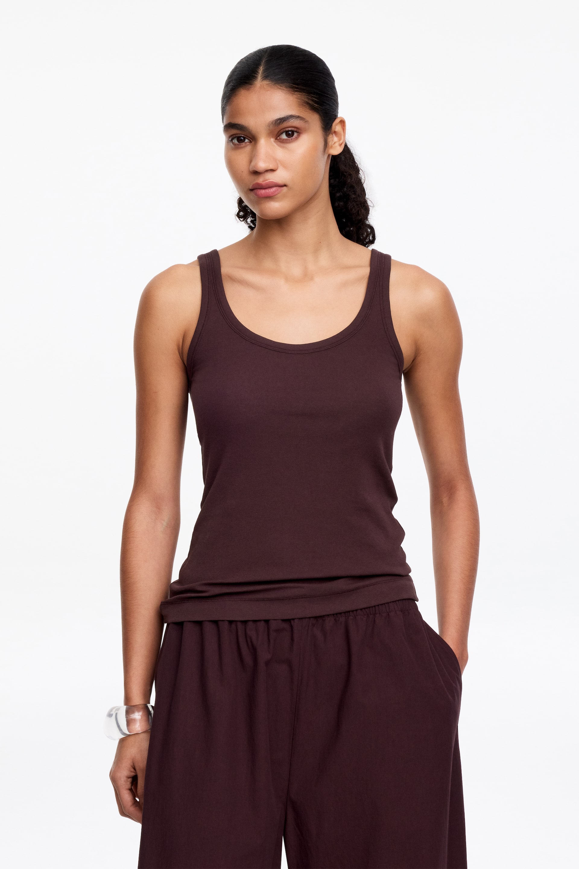 Zobacz większy obraz: Ciemnobrązowy top na ramiączkach prezentowany jest z przodu, z dekoltem w kształcie łódki i szerokimi ramiączkami. Przylega ściśle do biustu i tułowia, a jego dolna krawędź opiera się na pasku ciemnobrązowych spodni.