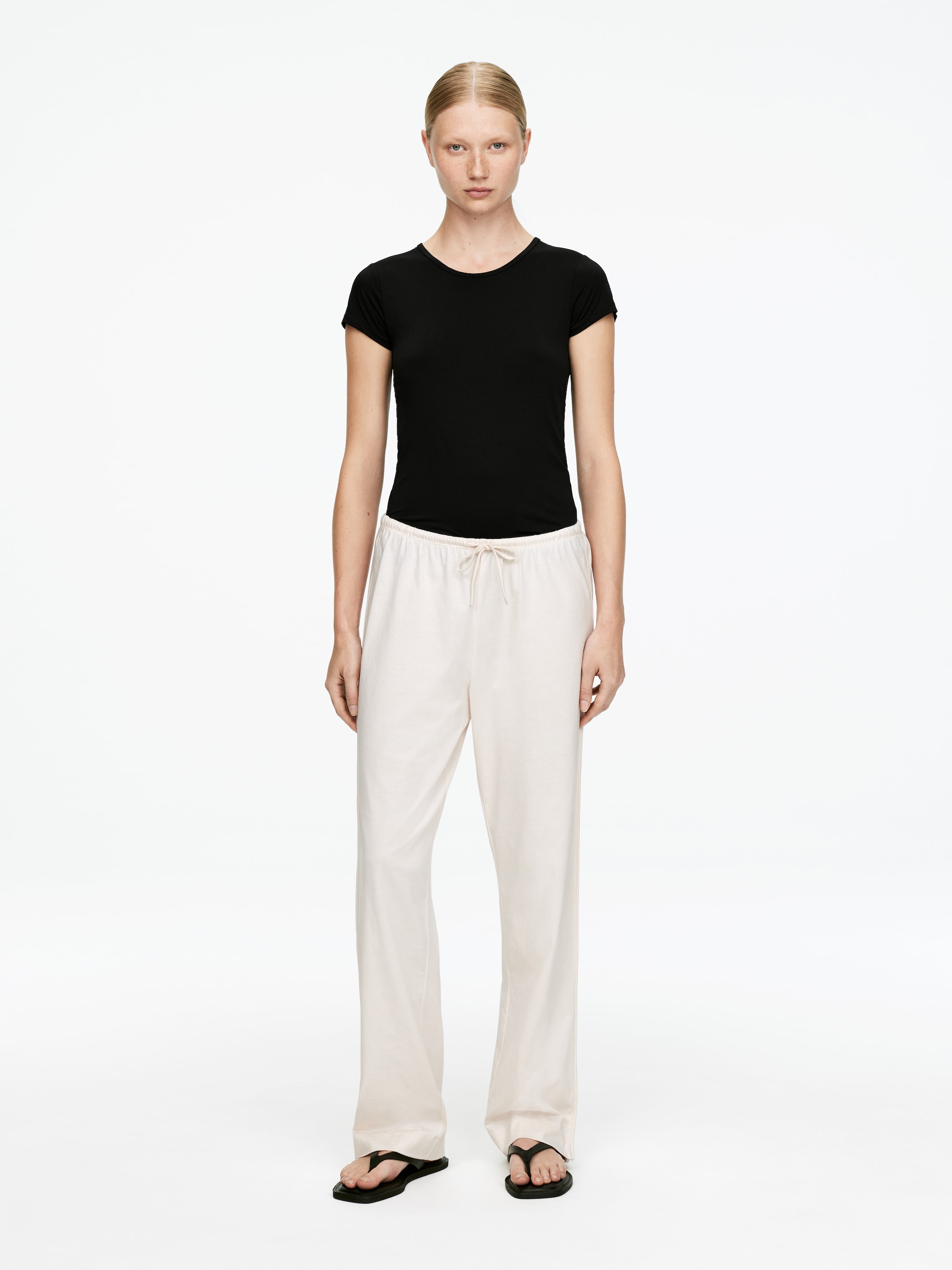 Grotere afbeelding bekijken: Een zwart, aansluitend T-shirt met korte mouwen en een ronde hals wordt gedragen, ingestopt in een licht beige, wijde broek met een zichtbare trekkoord taille en die lichtjes over zwarte teenslippers valt.