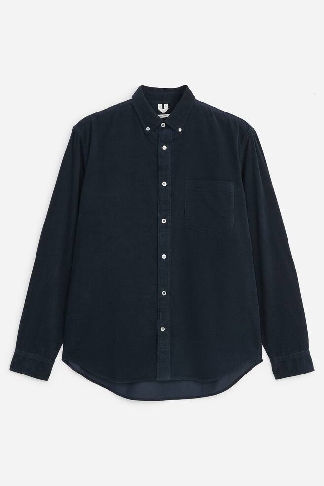 Una camicia in velluto a coste sottili blu navy scuro Ã¨ mostrata frontalmente, con classico colletto button-down, una singola tasca applicata sul petto e bottoni bianchi a contrasto. La camicia a maniche lunghe presenta polsini con bottoni e un orlo arrotondato. - 1