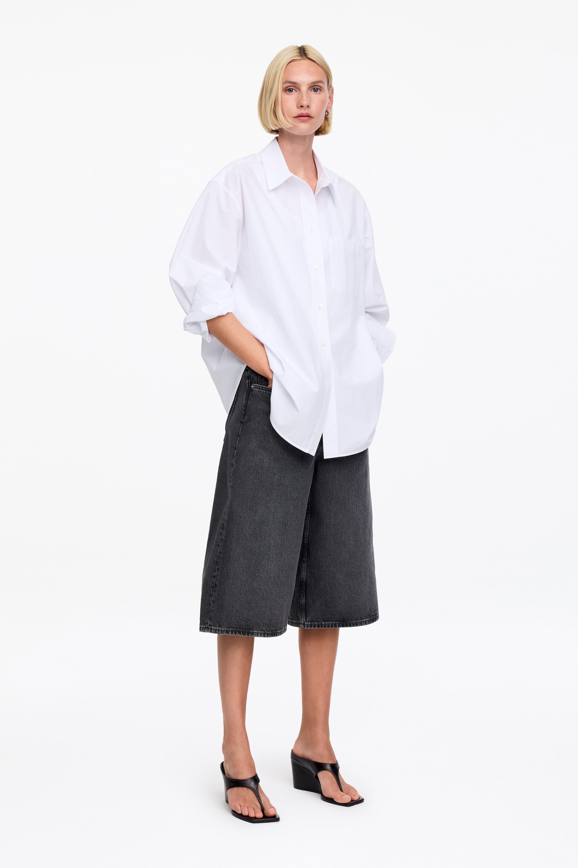 Ver imagen más grande: La modelo lleva puestos unos pantalones cortos de mezclilla de lavado oscuro y largo, con una silueta relajada hasta la rodilla. Está de pie con las manos en los bolsillos delanteros de los shorts, combinándolos con una camisa blanca de gran tamaño y sandalias negras de cuña.