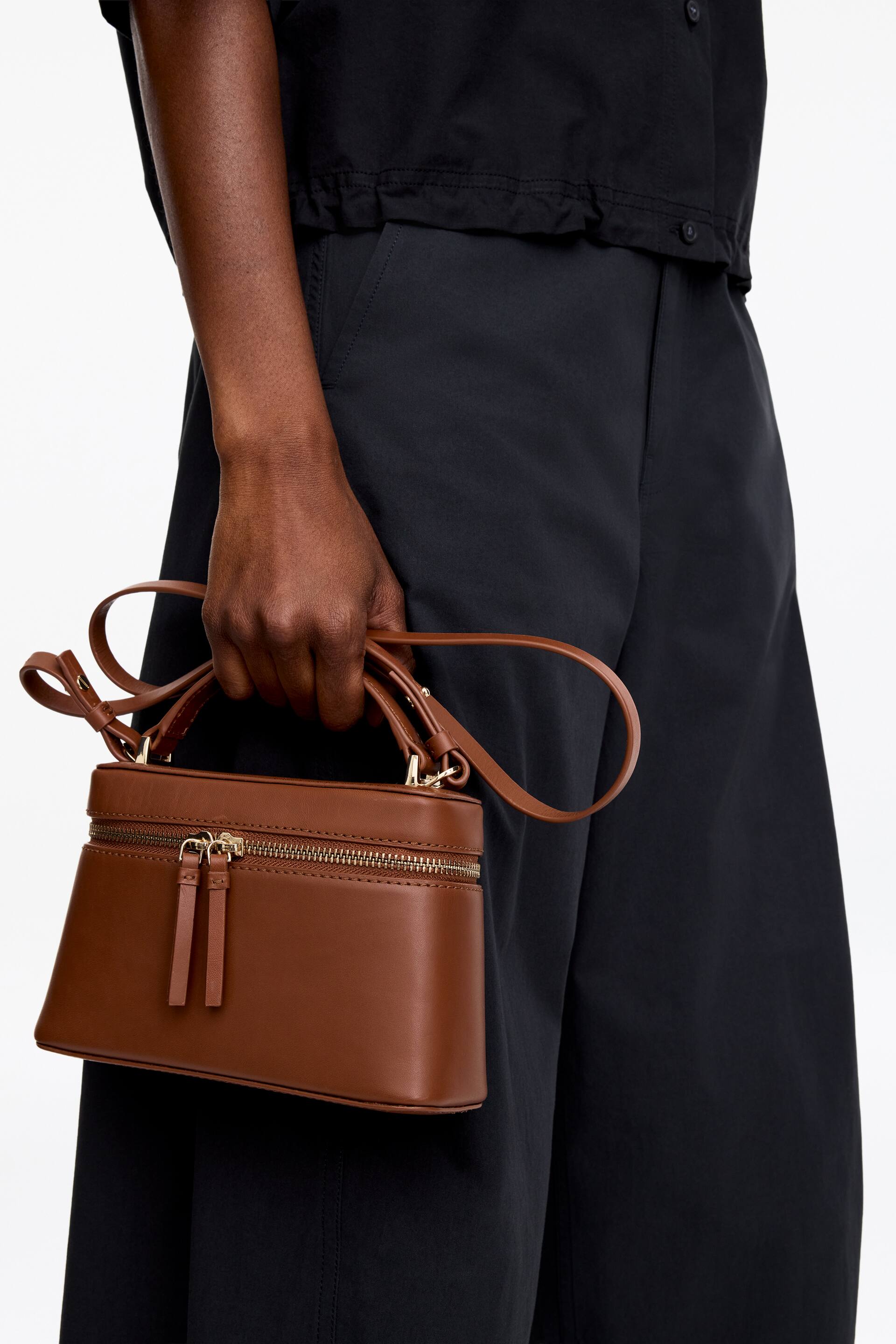 Visa större bild: En slät brun läder crossbody-väska med ett topphandtag, en avtagbar axelrem och en guldfärgad dragkedja med dubbla dragare, hålls av modellen.