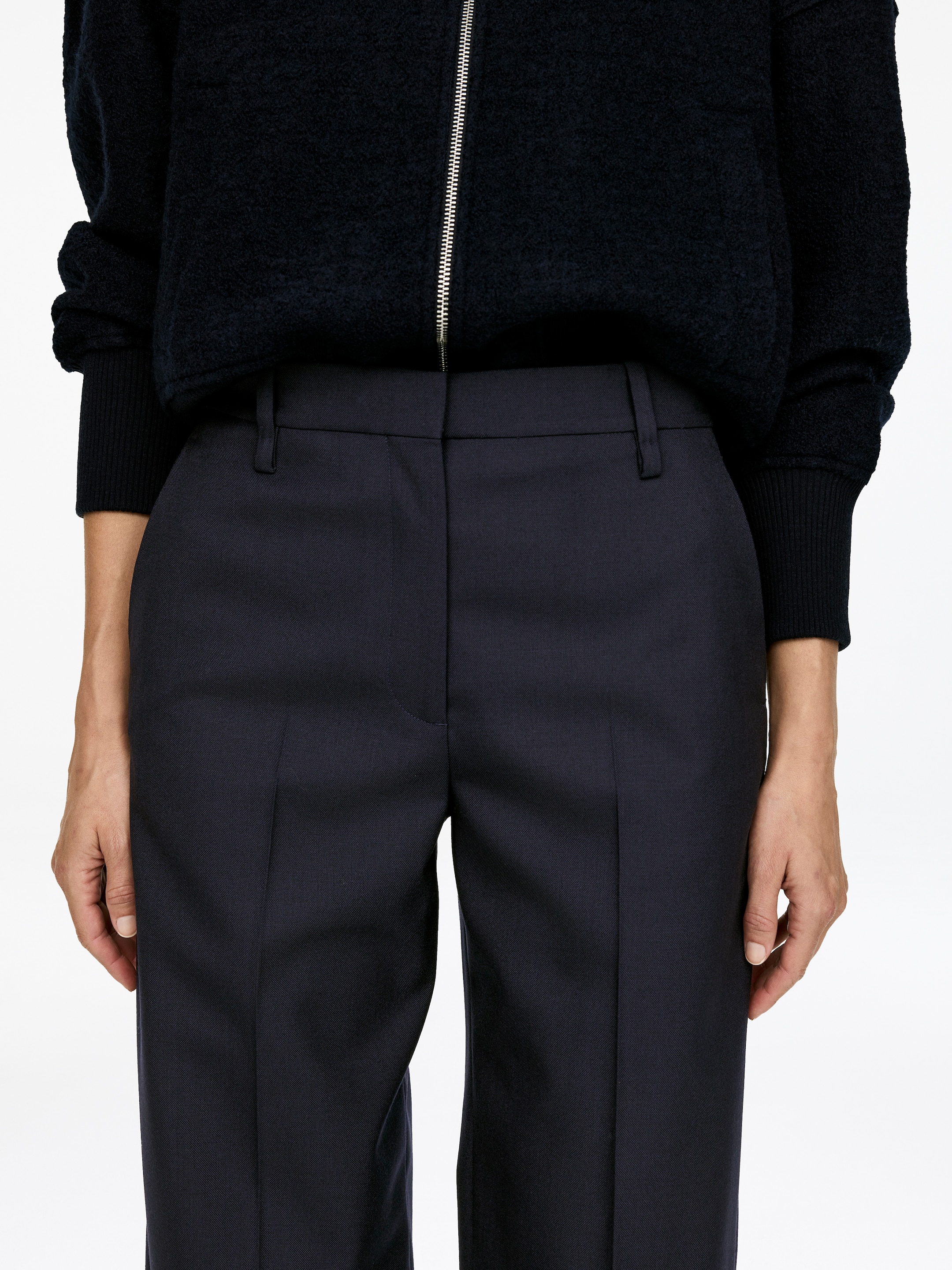 Agrandir l'image: Le mannequin porte un pantalon en laine sombre, doté d'une taille mi-haute et présentant des plis visibles à l'avant ainsi que des poches latérales. Une veste sombre et texturée est superposée au pantalon, couvrant la taille supérieure.