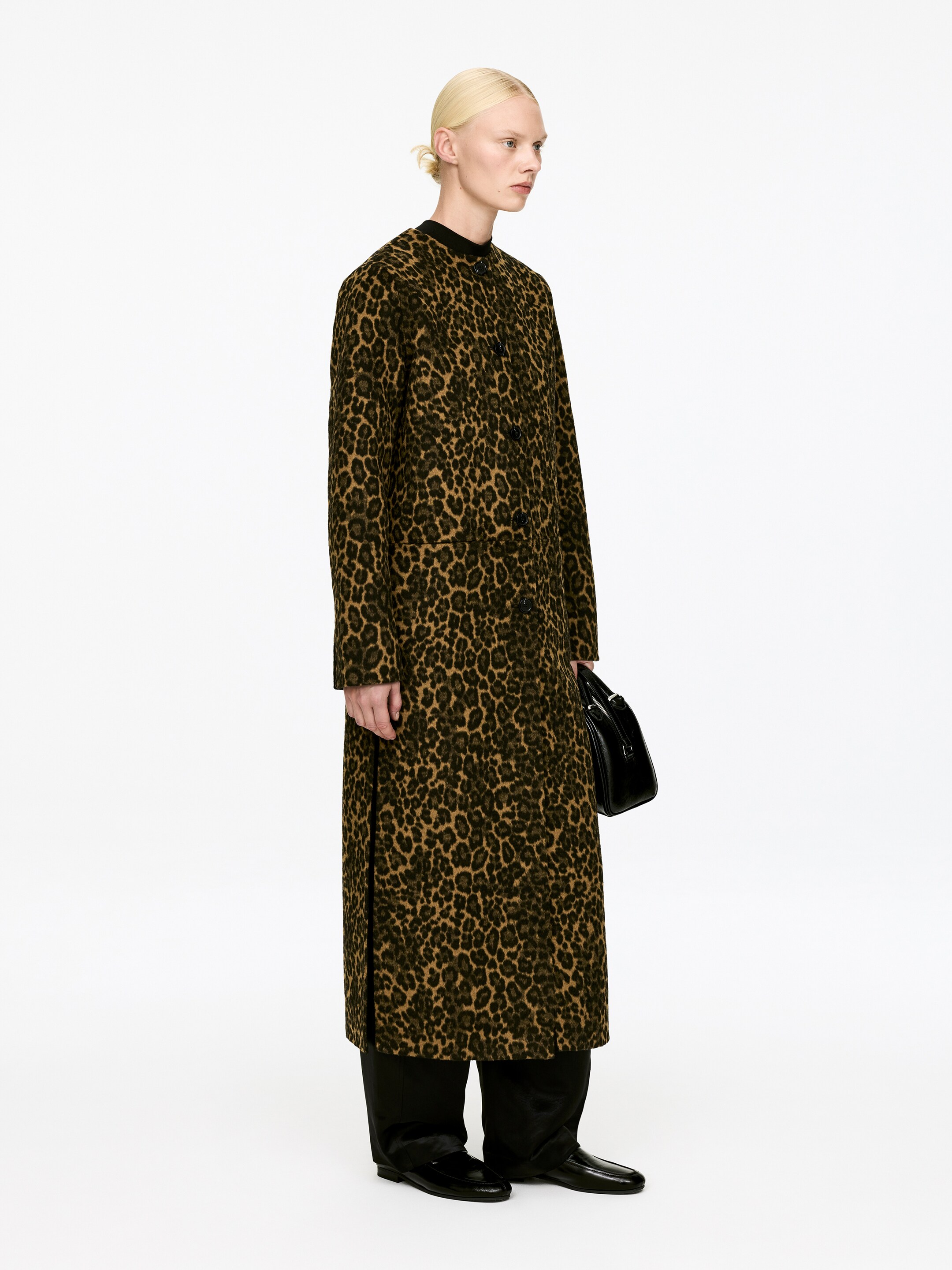 Vis større bilde: En lang leopardmønstret kåpe vises fra en sideprofil, vendt mot høyre. Den har en svart stående krage og en knappelukking foran, med en løs, rett silhuett som strekker seg til anklene.