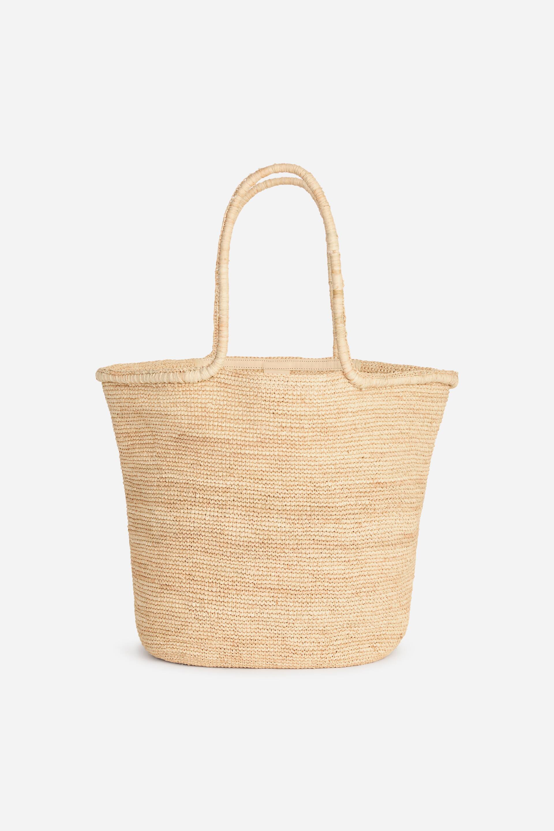 Agrandir l'image: Un sac cabas tissé en beige naturel clair est présenté de face, montrant une large ouverture et deux poignées arquées en tissu tissé s'étendant depuis le bord supérieur. Le sac arbore une texture tissée distincte et se rétrécit légèrement vers son fond plat.
