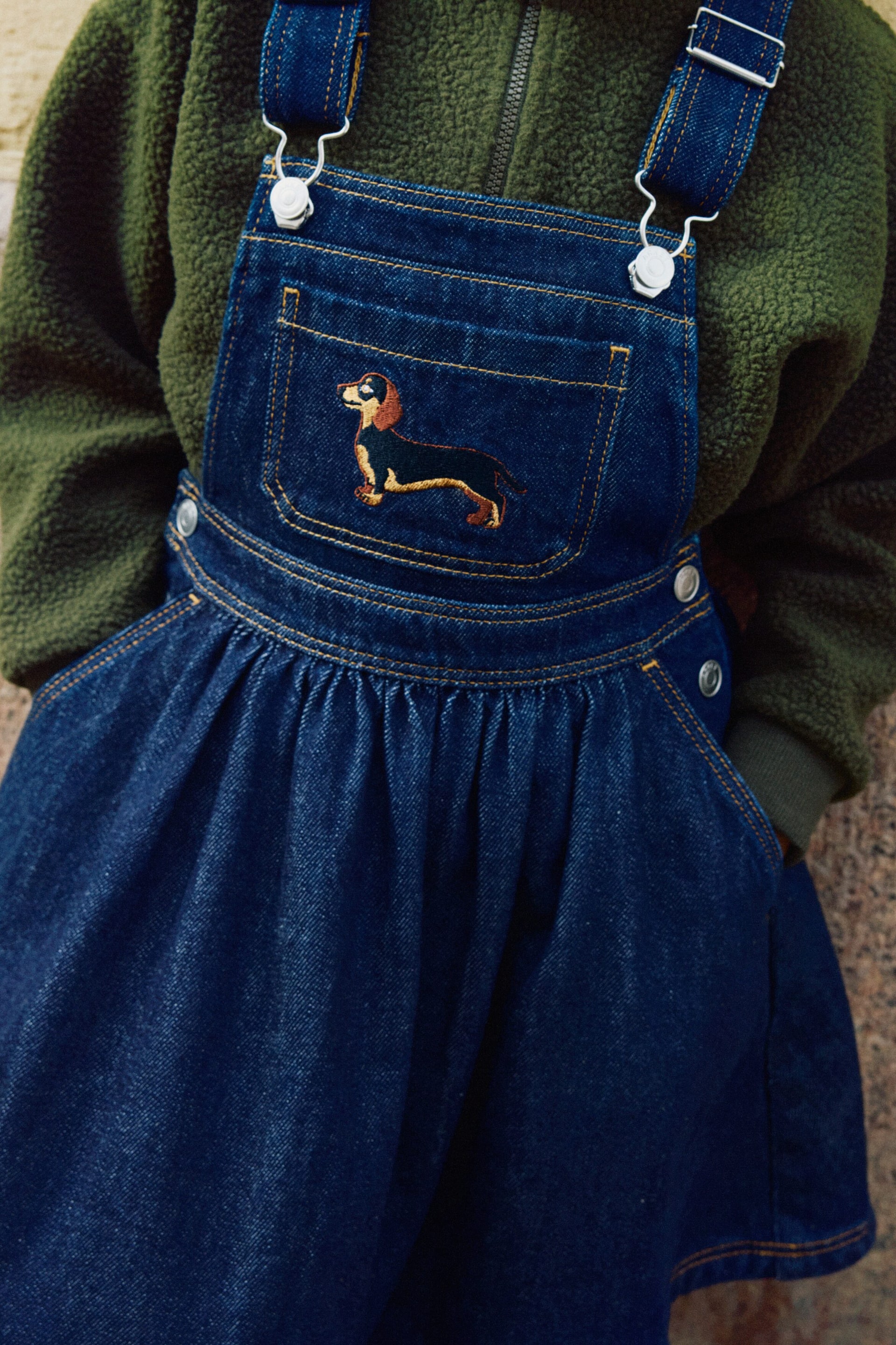 View larger image: A front view shows a dark blue denim pinafore dress layered over a dark green pile half-zip sweater. The pinafore dress features adjustable shoulder straps with metal buckles, side popper buttons, and a bib pocket embroidered with a dachshund, while the sweater has a raised collar and ribbed cuffs.
