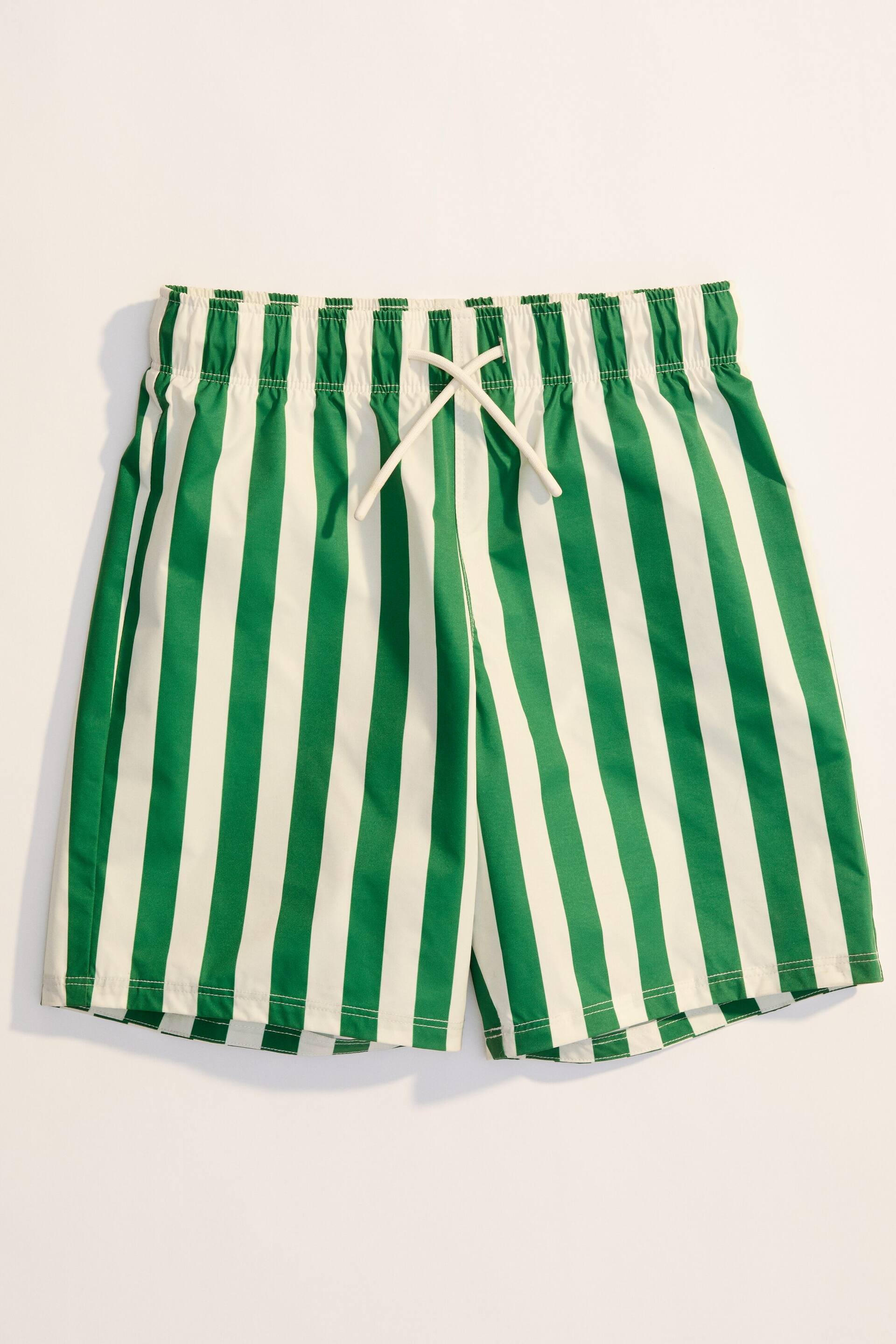 Größeres Bild ansehen: Badeshorts zeigen ein Muster aus kräftigen grünen und weißen vertikalen Streifen. Sie verfügen über einen elastischen Bund mit einer weißen Kordel, die vorne gebunden ist, und sind mit etwas längeren Beinen gestaltet.