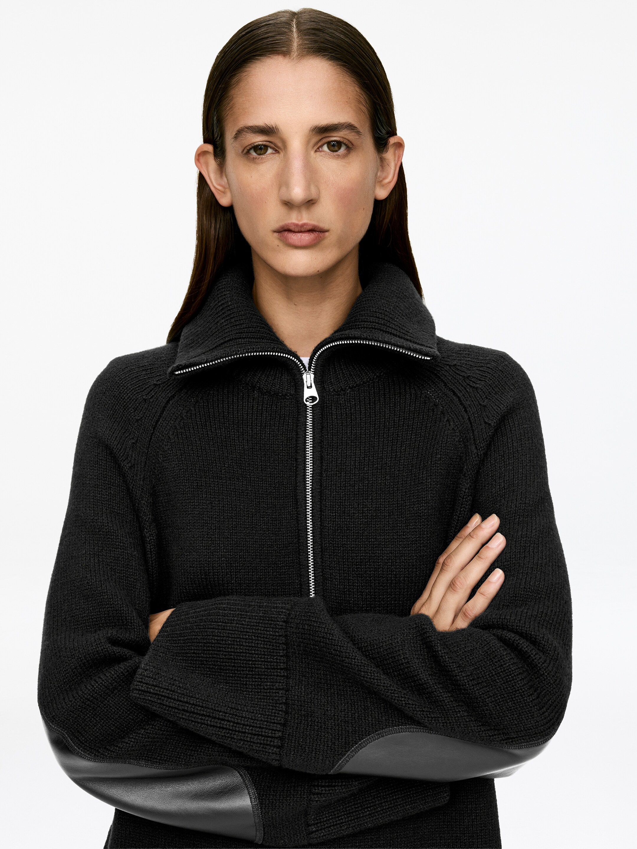 Visualizza immagine più grande: Un maglione scuro a mezza zip con vestibilità rilassata, caratterizzato da una zip argentata parzialmente aperta al colletto e toppe di pelle scura sui gomiti, è indossato dalla modella. La modella incrocia le braccia, enfatizzando visivamente le toppe sui gomiti.