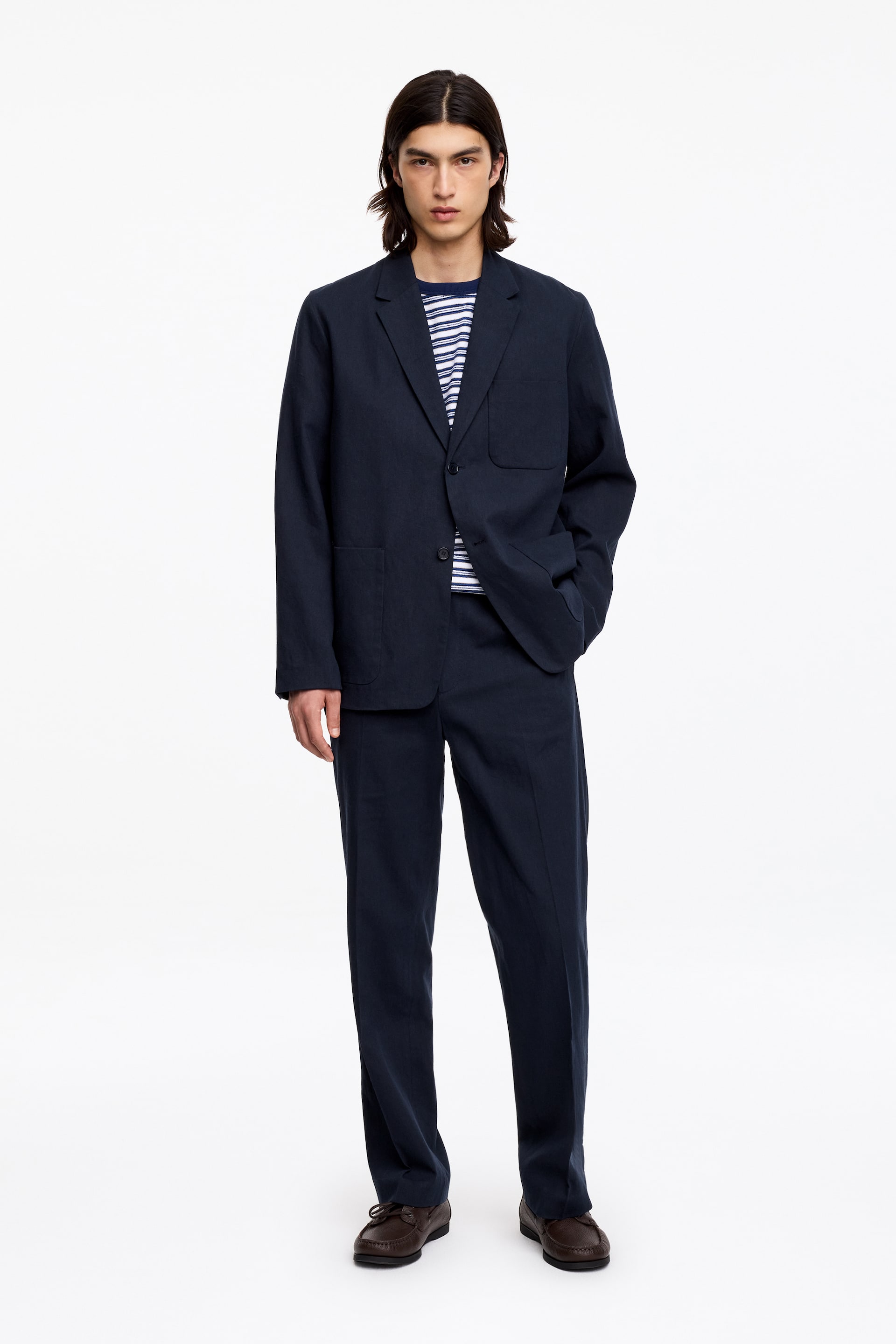 Agrandir l'image: Un costume deux pièces bleu marine est présenté, comprenant une veste à simple boutonnage avec des revers crantés, une fermeture à deux boutons avec un bouton fermé, et trois poches plaquées. Un t-shirt rayé est visible sous la veste, tandis que le pantalon assorti offre une coupe droite et décontractée.