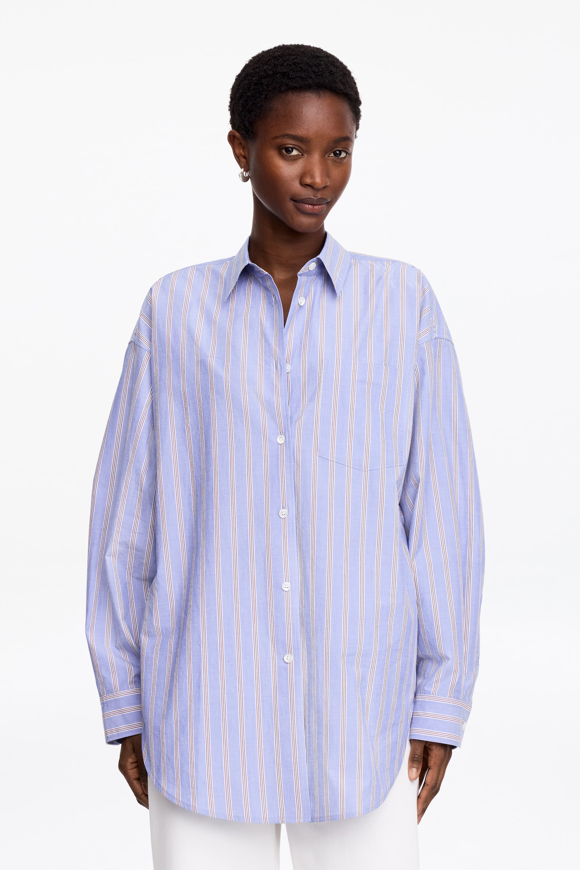 Ver imagem maior: Uma camisa azul clara oversized apresenta finas riscas verticais brancas e vermelhas. É desenhada com uma gola pontiaguda, uma frente com botões brancos, mangas compridas com punhos abotoados, um bolso de remendo no peito esquerdo e uma bainha arredondada, voltada para a frente.