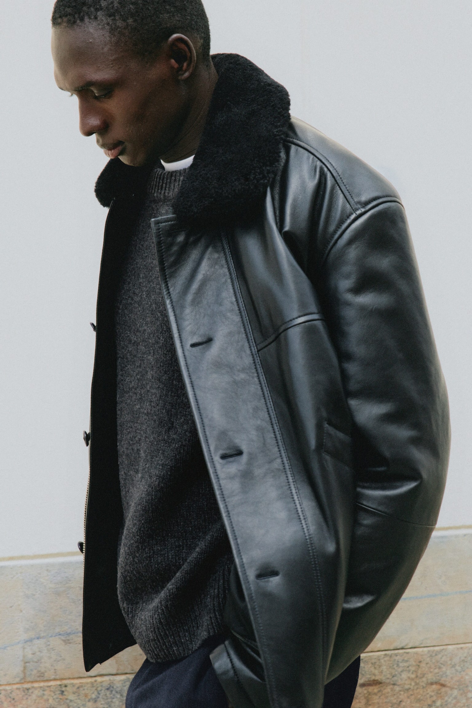Eine schwarze Leder-Fliegerjacke wird über einem dunkelgrauen Strickpullover getragen und betont ihren weichen schwarzen Shearling-Kragen sowie die lockere Passform. Die Jacke ist in einer Dreiviertelansicht nach links ausgerichtet und verfügt über eine glatte Lederoberfläche sowie eine geknöpfte Frontleiste. - 1