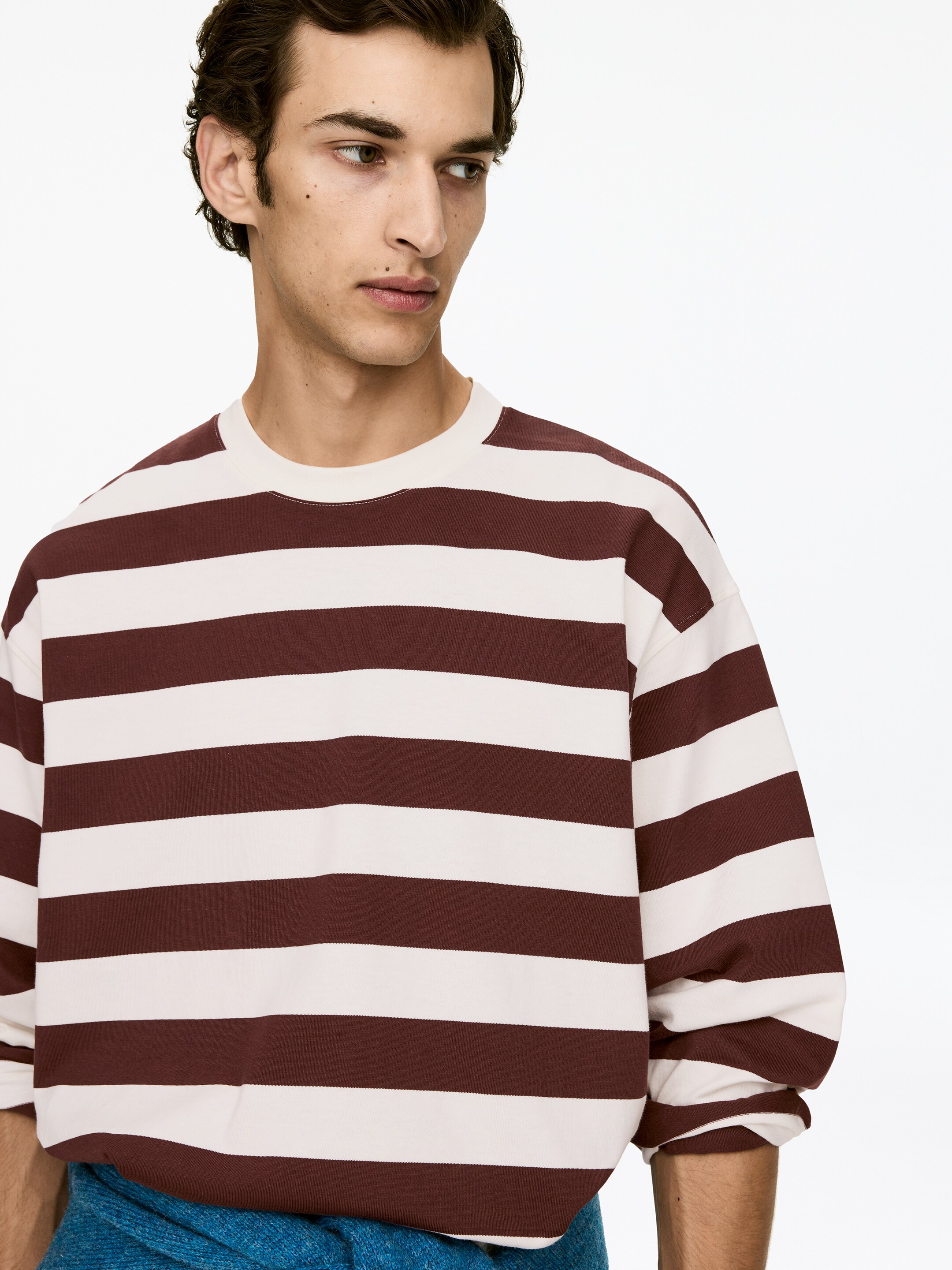 Agrandir l'image: La chemise à manches longues présente des rayures horizontales larges en bordeaux et blanc cassé, avec un col rond blanc cassé. Elle affiche une coupe décontractée avec des manches longues légèrement surdimensionnées, et son ourlet repose sur un vêtement bleu.