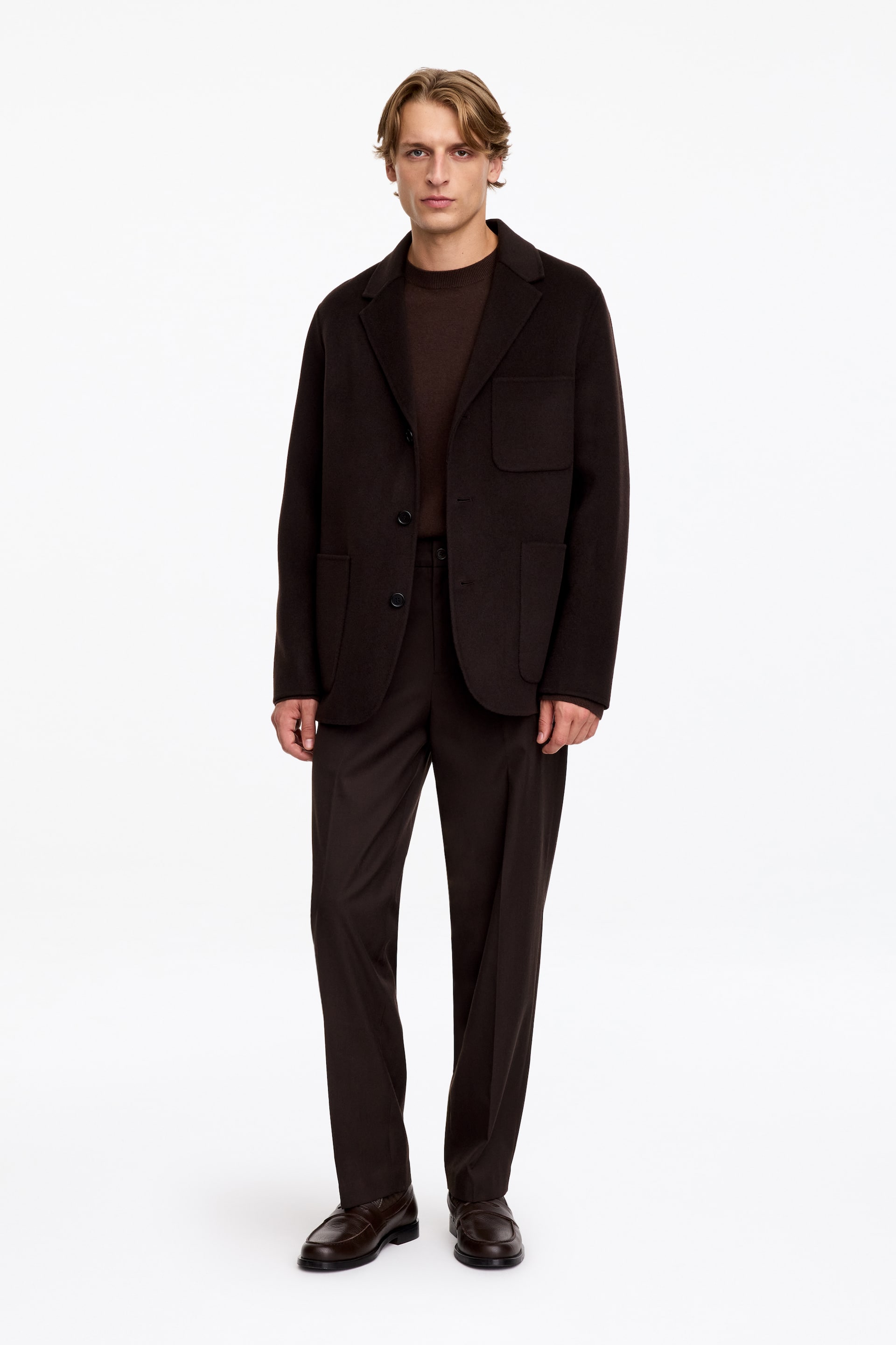 Visa större bild: Dessa mörkbruna byxor med avslappnad passform och pressad mittpress bärs av modellen. Byxorna stylas med en matchande mörkbrun blazer, en mörkbrun rundhalsad tröja och mörka loafers i läder.