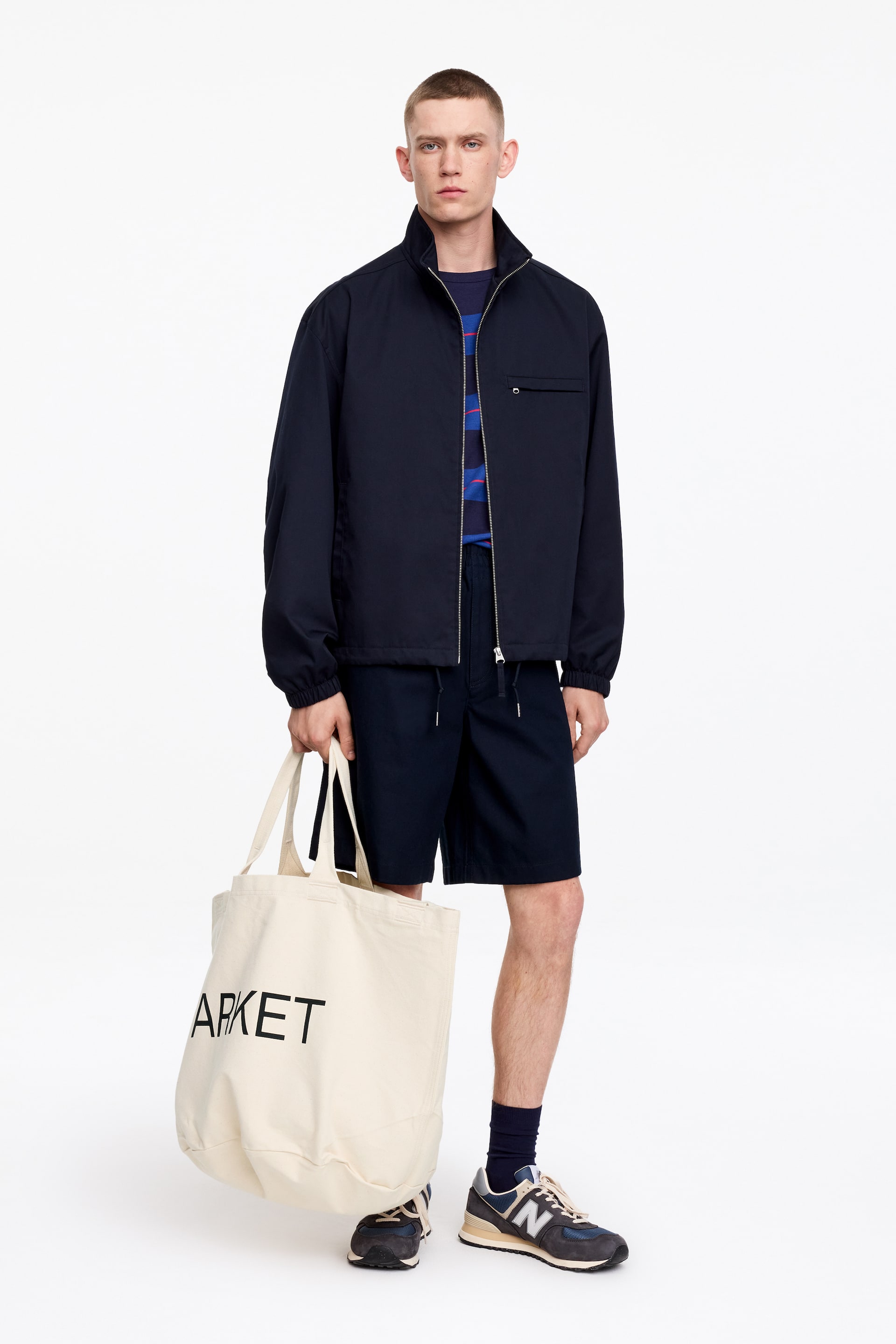 Visualizza immagine più grande: Una giacca blu navy con zip completa, caratterizzata da un colletto alla coreana, una tasca sul petto con patta e polsini e orlo elasticizzati, è mostrata su un modello in piedi, leggermente rivolto a destra. È indossata sopra una t-shirt con motivo blu e abbinata a pantaloncini con coulisse blu navy, calzini blu scuro e sneakers blu e grigie. Una borsa tote in tela color crema con la scritta "ARKET" in lettere nere viene tenuta in mano.