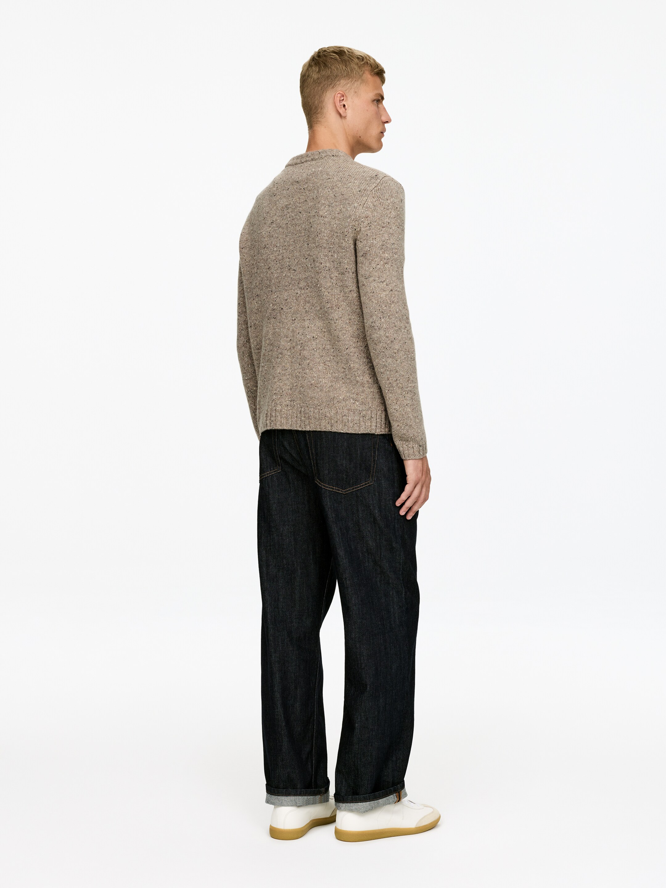 Se større billede: En beige meleret crew-neck sweater bæres, set fra bagsiden til højre, med ribstrikkede manchetter og søm. Den er stylet med mørke vasket, bredbenede jeans, som er rullet op ved anklen for at afsløre et lysere indre, og som hviler på hvide sneakers med gummisåler.