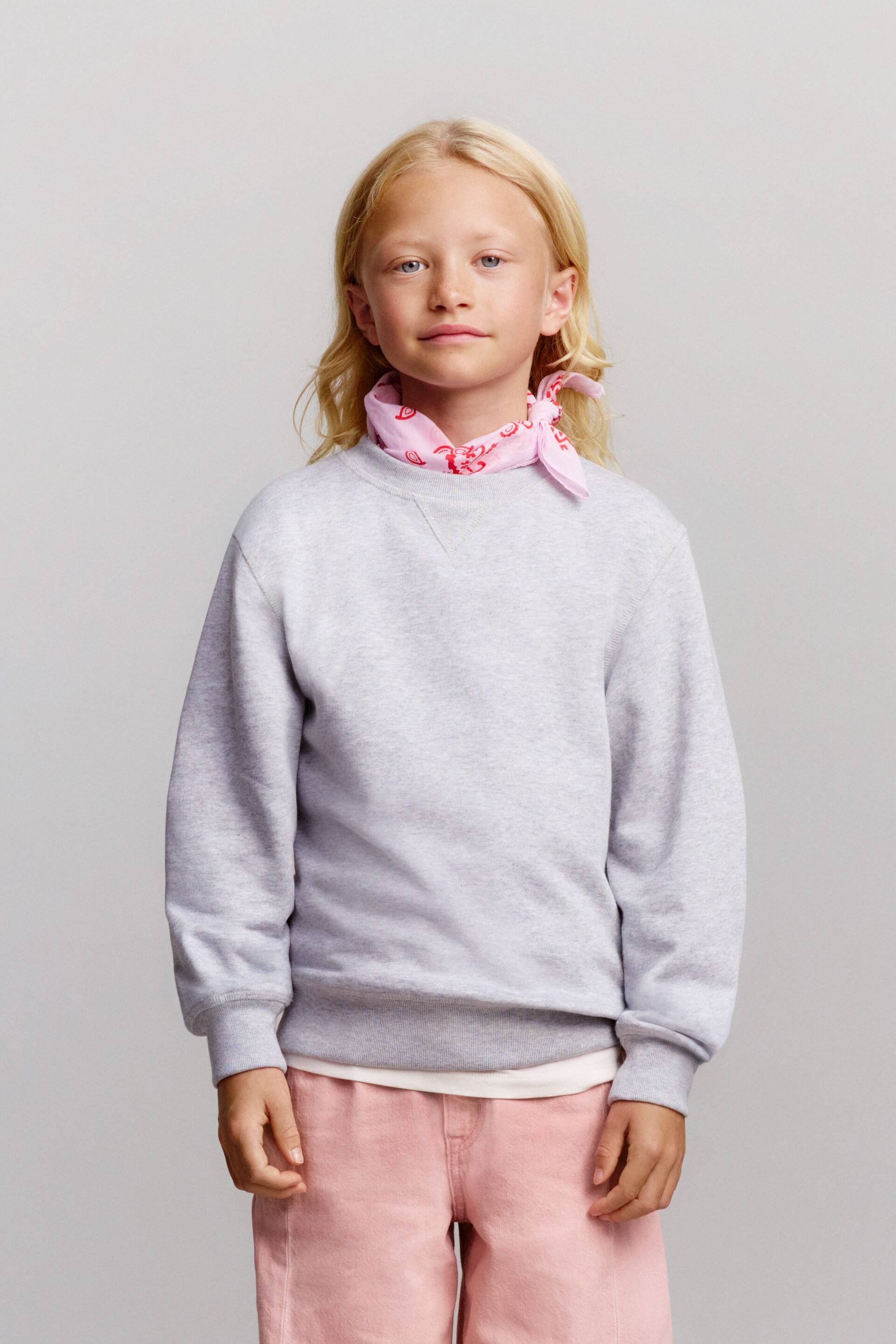 Agrandir l'image: Un sweat-shirt à col rond gris clair avec finitions côtelées et détail en V est présenté de face, stylé avec un jean barrel coupe droite rose pâle et un bandana rose assorti à motif cachemire noué autour du cou.