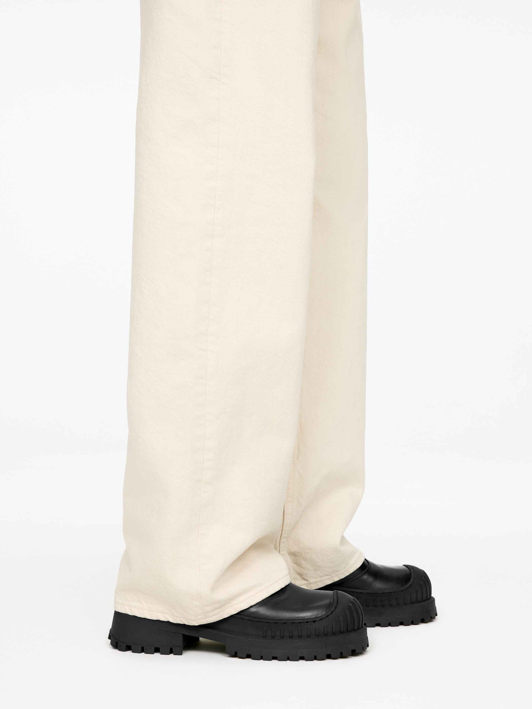 Grotere afbeelding bekijken: Lichtbeige broek met wijde pijpen valt over zwarte, grove schoenen, getoond vanuit een zijaanzicht. De broek heeft een ontspannen pasvorm, die over de bovenkant van de schoenen valt, terwijl de schoenen dikke, lug-zolen en gestructureerde teennaden hebben.