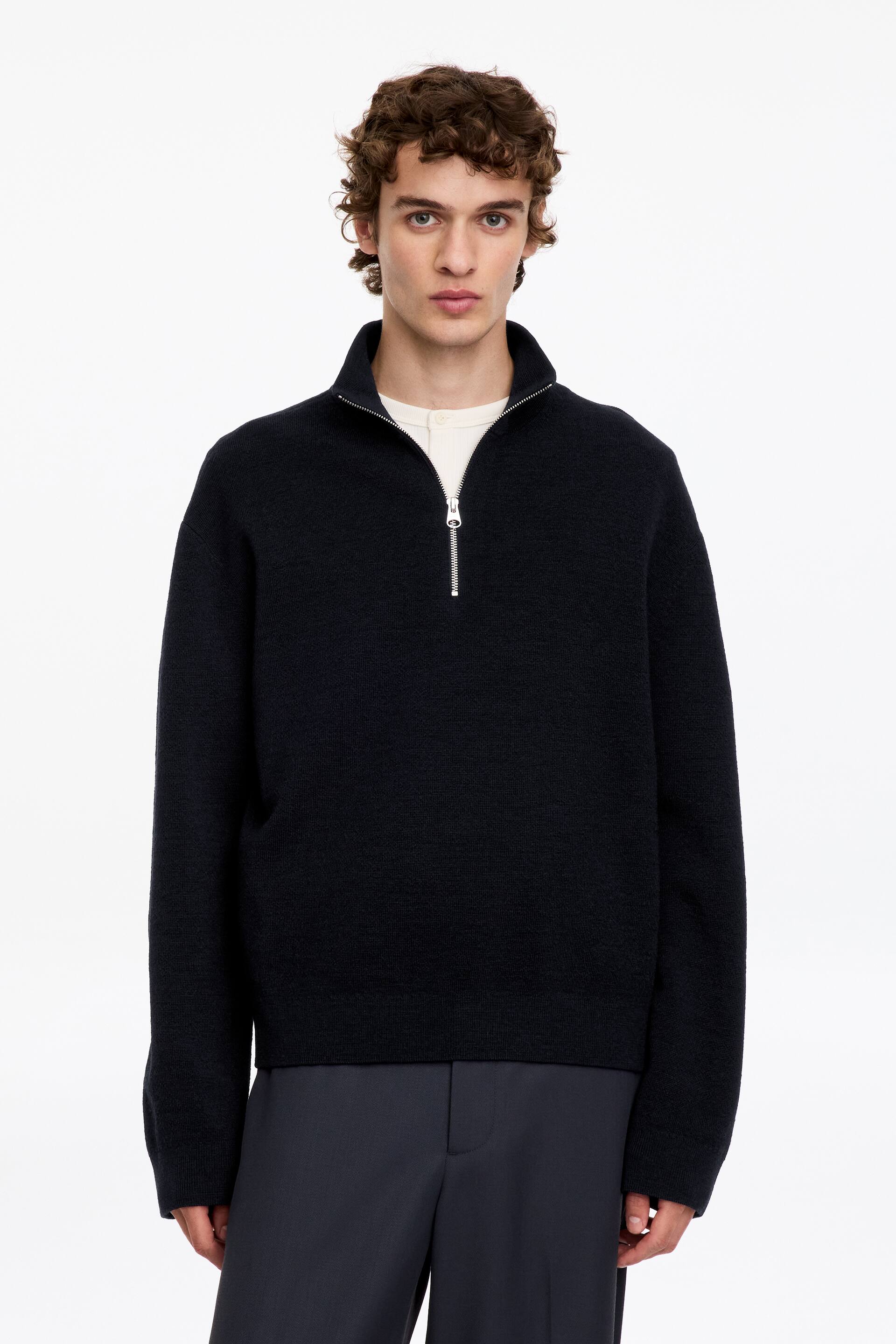 View larger image: The model is wearing a black knitted half-zip jumper, featuring a silver-tone metal zip and a high collar. The jumper is layered over a cream shirt and styled with dark grey trousers.