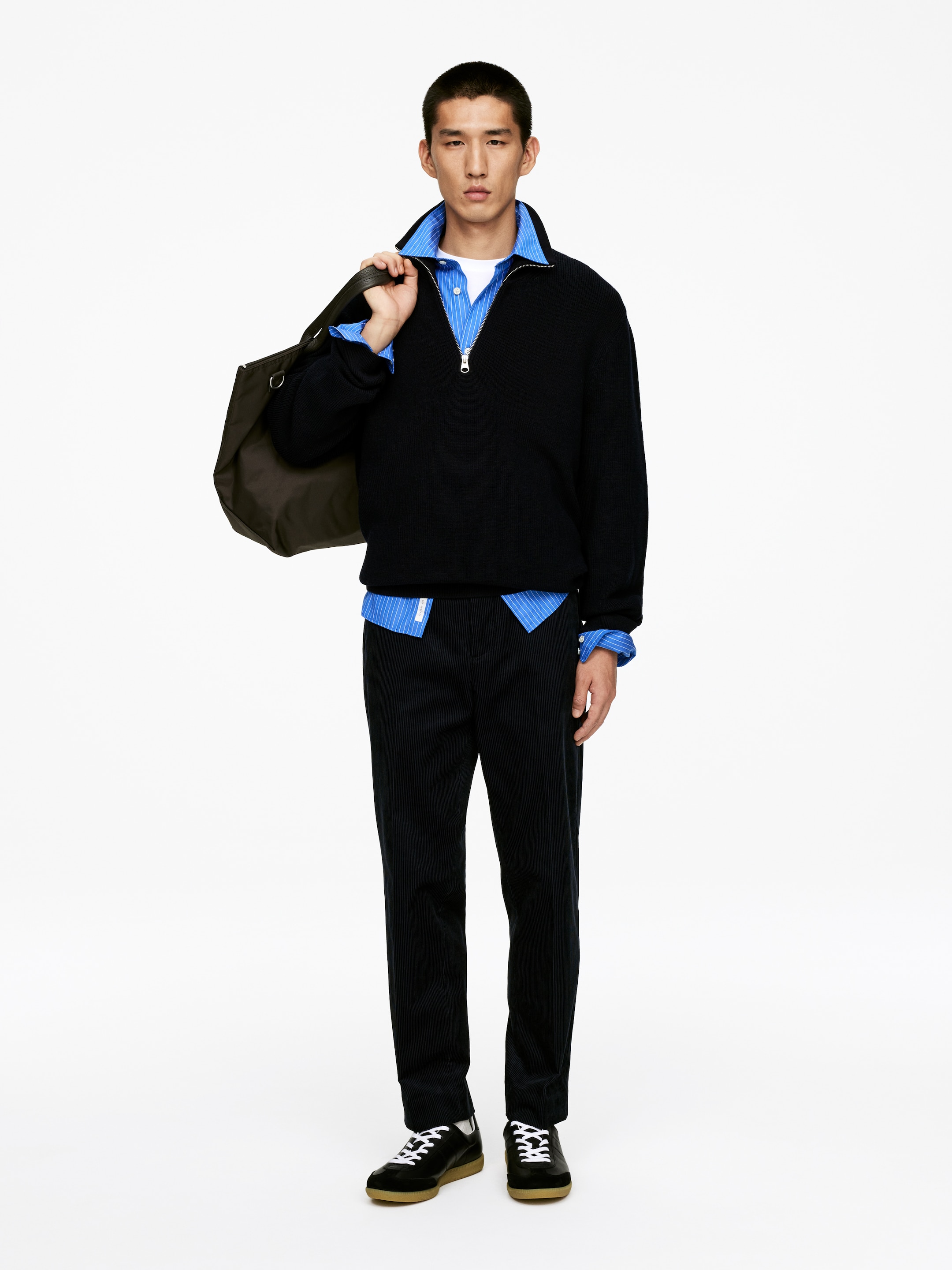 Visualizza immagine più grande: Un maglione nero con mezza zip, caratterizzato da un colletto alto, polsini a coste e un orlo a coste, è stratificato su una camicia a righe blu e bianca e una t-shirt bianca. Pantaloni in velluto a coste scuri con una vestibilità dritta completano il look, insieme a sneakers nere che presentano lacci bianchi e suole in gomma. Una borsa a tracolla scura è appoggiata sulla spalla sinistra.