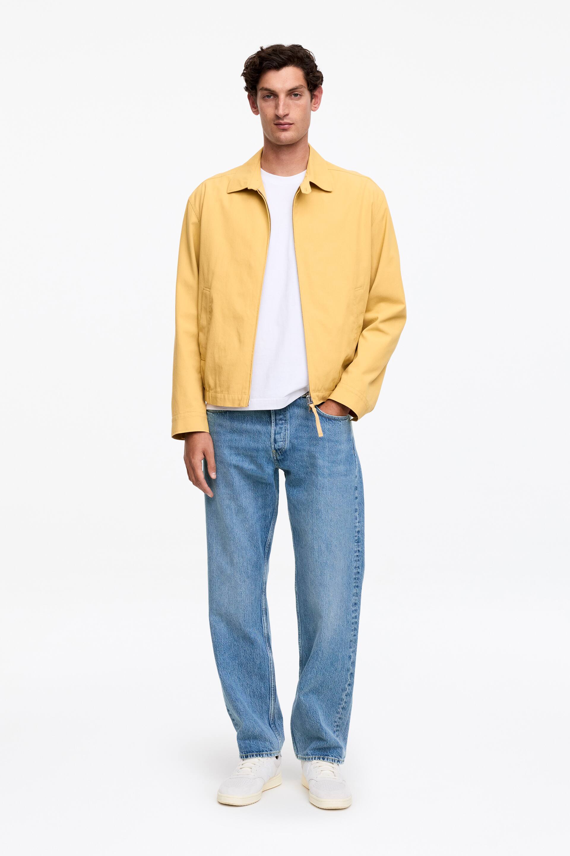 Ingrandisci l'immagine: Una giacca casual gialla è presentata da un'angolazione frontale, con un colletto a punta, una chiusura centrale con zip visibile e un orlo con fascia in vita. Le maniche lunghe si estendono dalle spalle e la giacca è indossata sopra una t-shirt bianca, con una mano appoggiata in una tasca laterale.