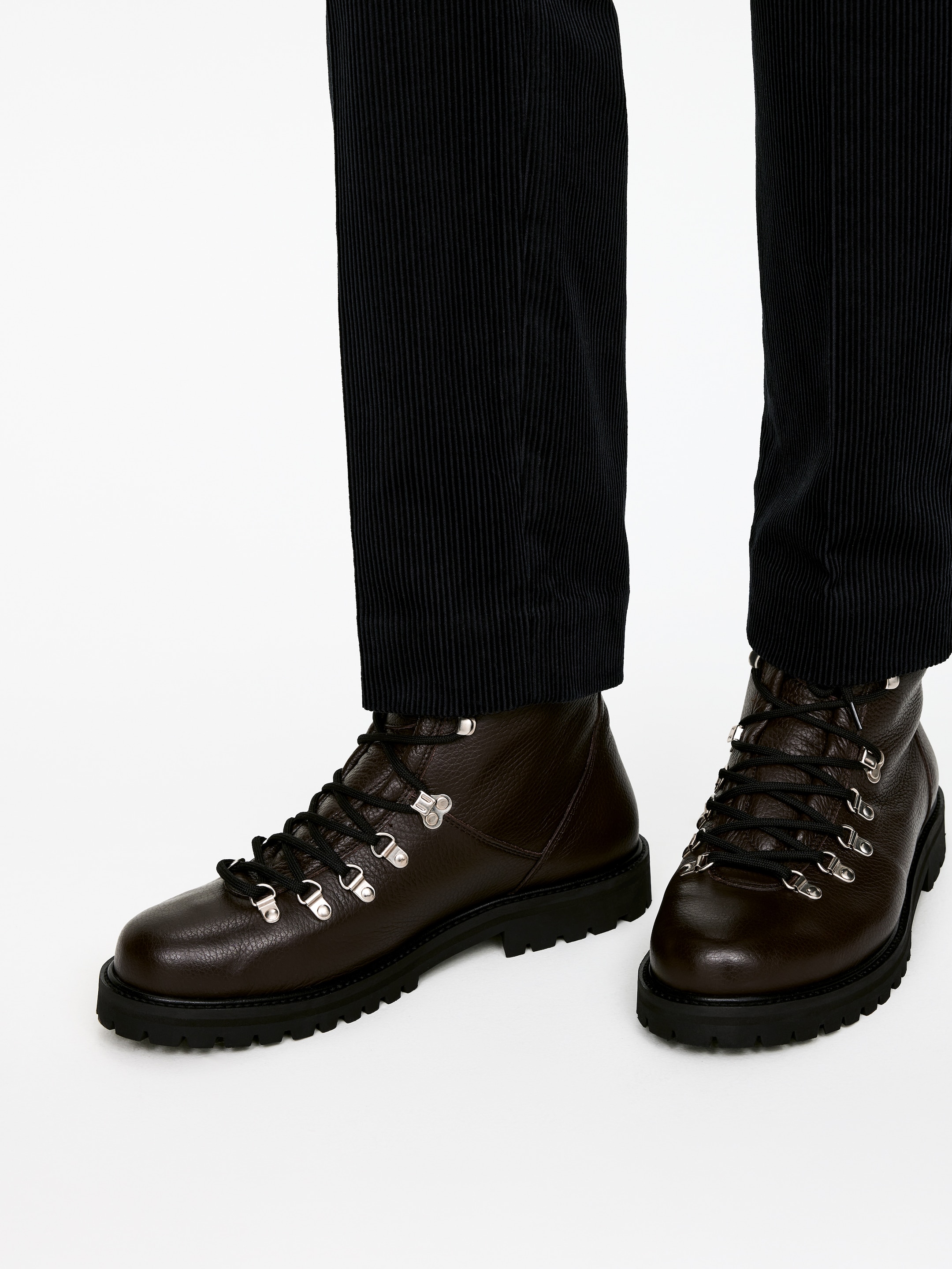 Ver imagem maior: Botas de montanha em couro castanho escuro com textura estão a ser usadas, com cordões pretos passados por ilhós em forma de D em prata e ganchos de fecho rápido. Estão assentes numa sola negra grossa com tacos e estão parcialmente cobertas por calças caneladas escuras.