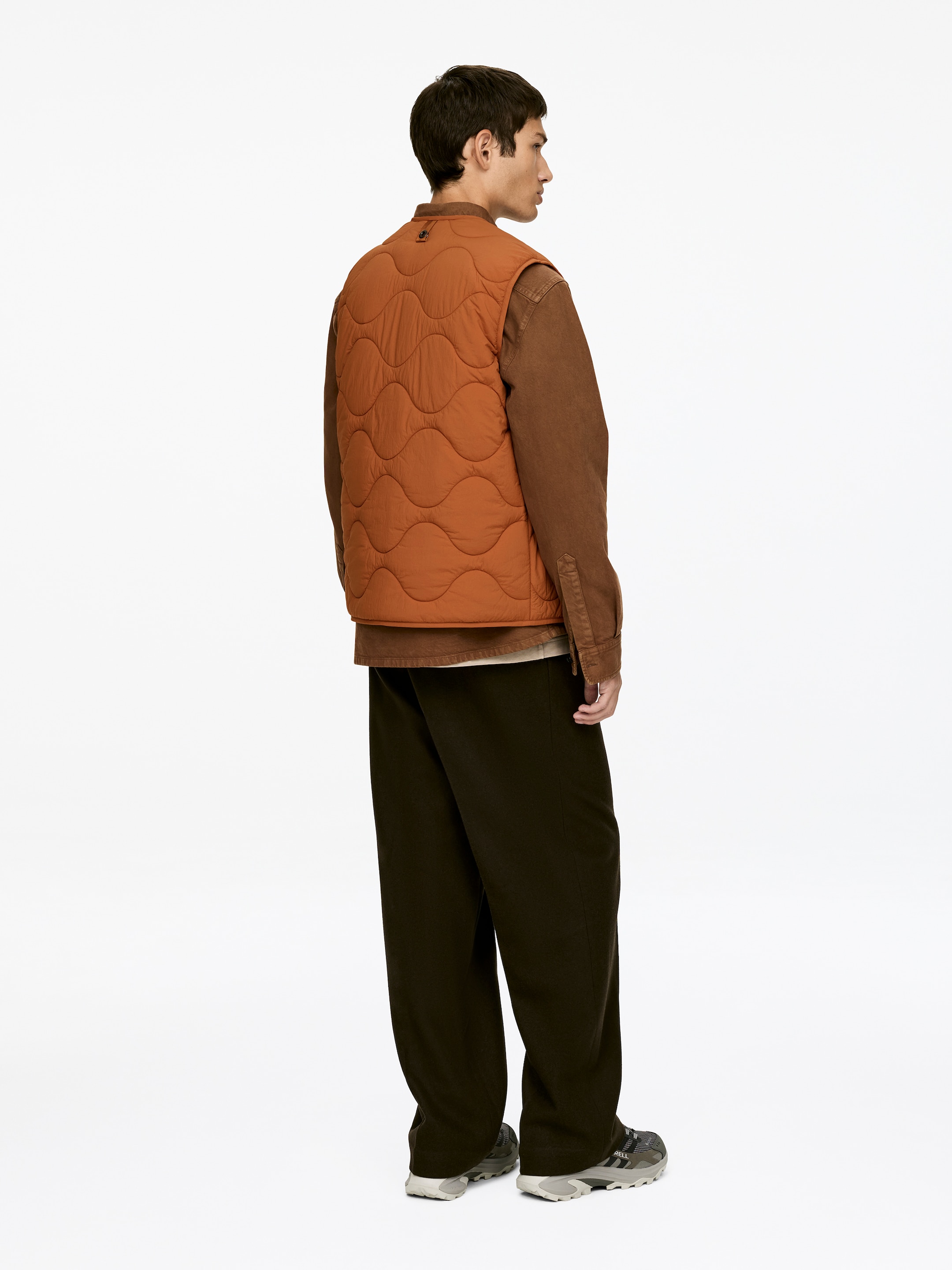 Agrandir l'image: Un gilet matelassé d'un chaleureux brun orangé est présenté sous un angle arrière droit, affichant un motif ondulé sur son dos et un col montant bas. Il est porté superposé à une chemise marron à manches longues et à col, tandis que des pantalons marron foncé et amples couvrent les jambes, et des baskets grises et noires apparaissent sur les pieds.