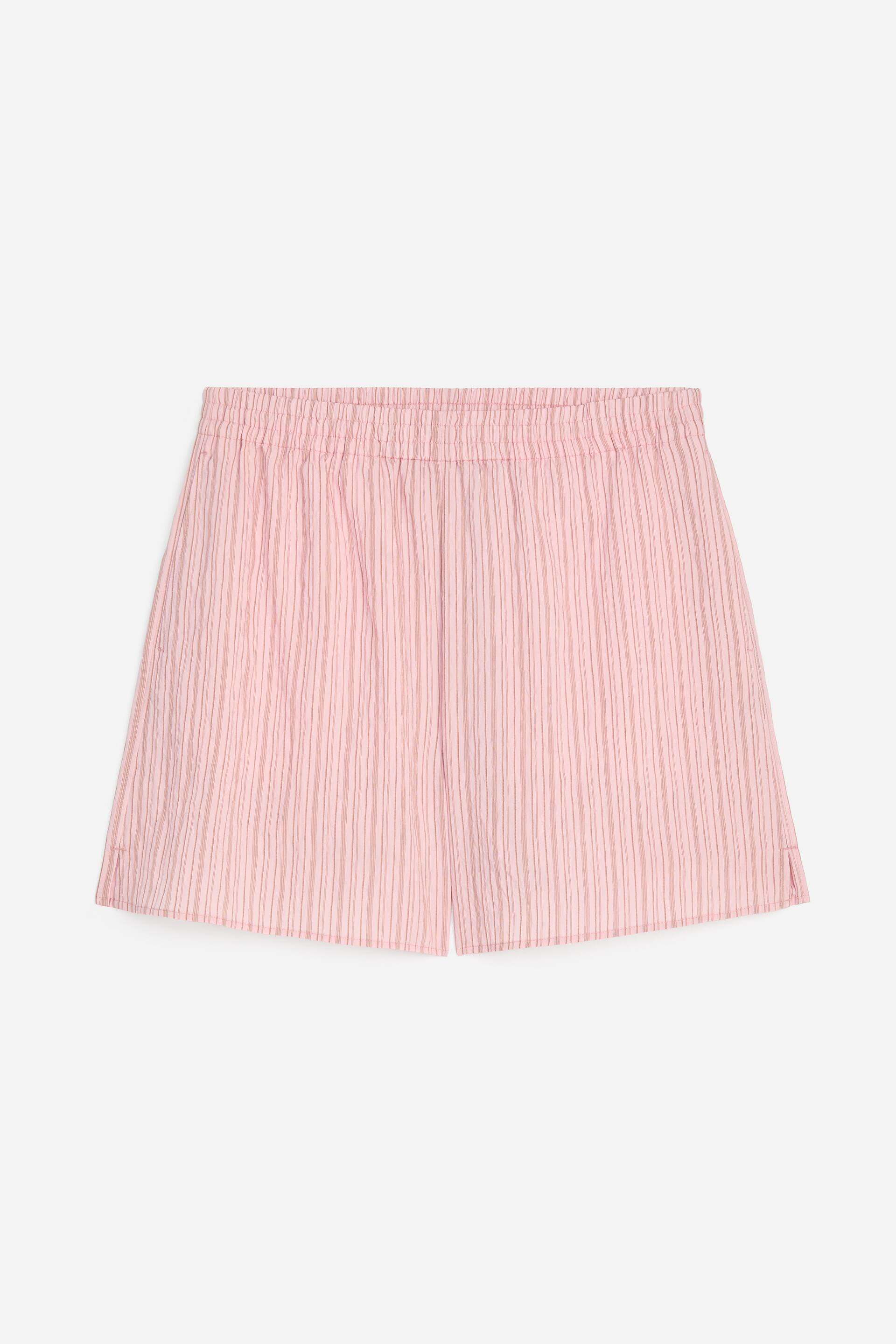Visualizza immagine più grande: Pantaloncini rosa mostrano una vista frontale, caratterizzati da sottili righe verticali e una vita elasticizzata e arricciata. L'orlo dritto presenta piccoli spacchi laterali.