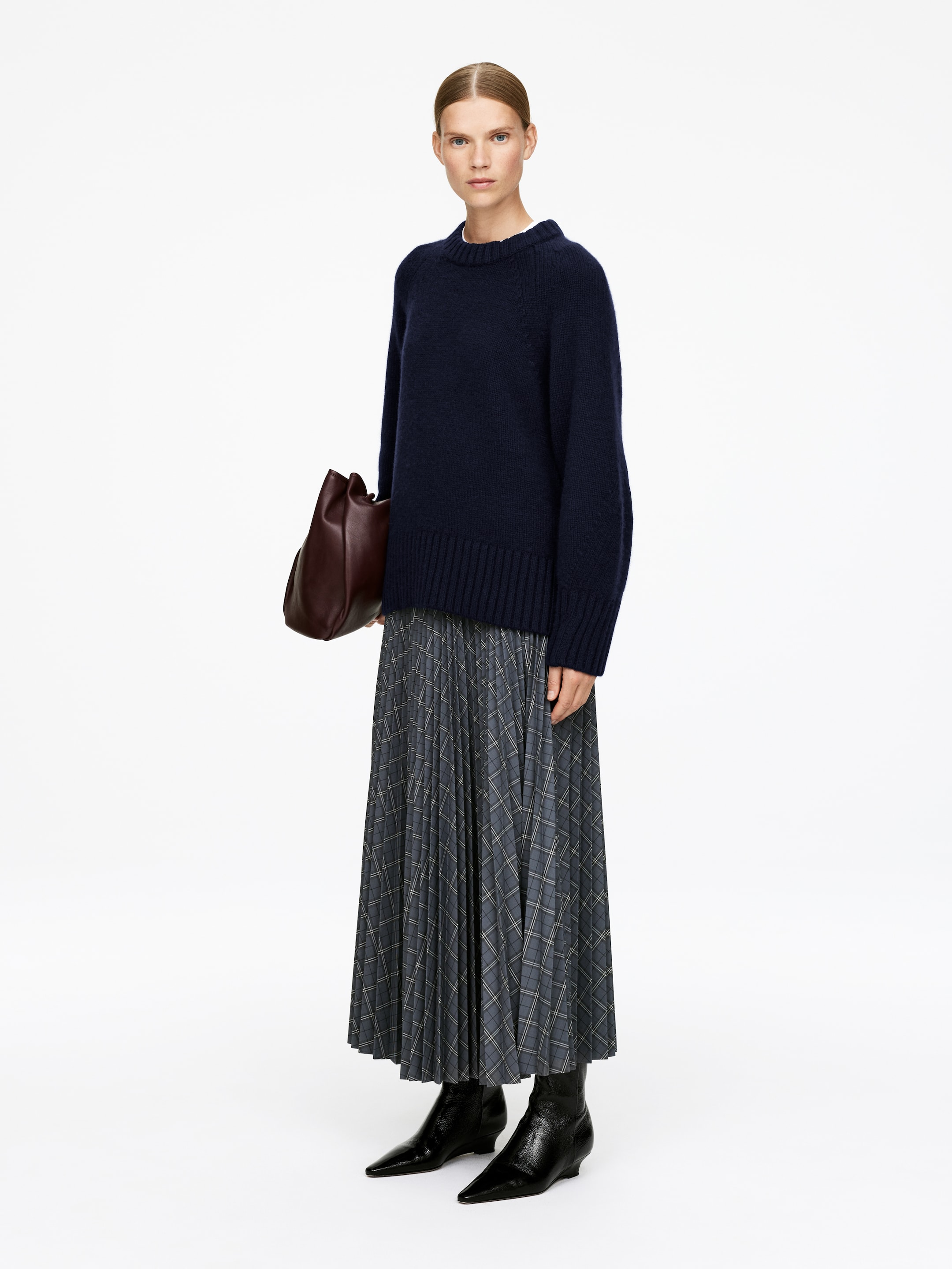 Visualizza immagine più grande: Un maglione girocollo blu navy, caratterizzato da una vestibilità rilassata e polsini e orlo a coste larghe, è indossato con una gonna grigia a pieghe a tutta lunghezza che presenta un motivo a quadri. Stivali neri a punta e una borsa in morbida pelle bordeaux completano l'ensemble stilizzato.
