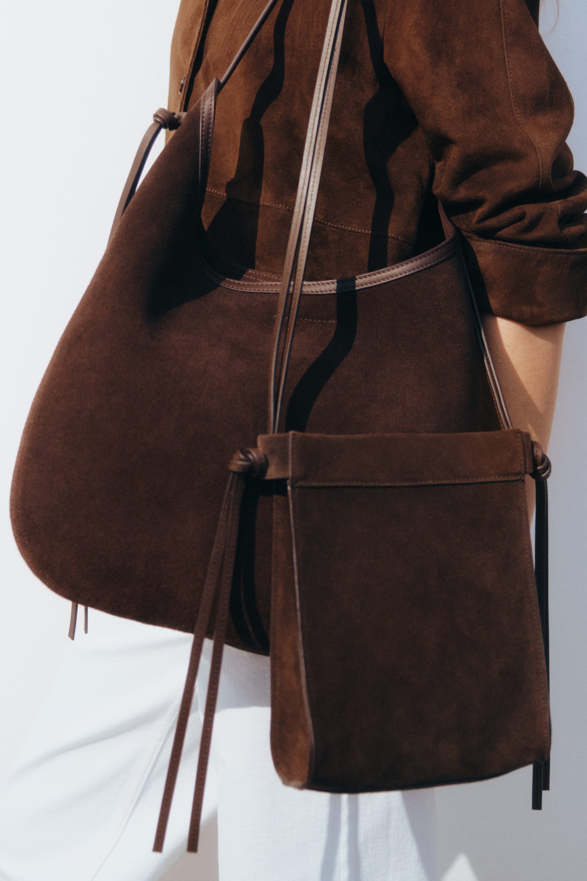 Agrandir l'image: Une chemise en daim marron, avec des empiècements visibles et des poignets boutonnés retroussés, est portée, avec deux sacs en daim marron superposés par-dessus. Un grand sac à bandoulière arrondi est exposé à l'AVANT, sa bandoulière ajustable et ses fines lanières latérales visibles, tandis qu'un sac bandoulière plus petit, de forme rectangulaire, avec des détails en cuir fin, est porté à l'AVANT, positionné devant le sac plus grand.