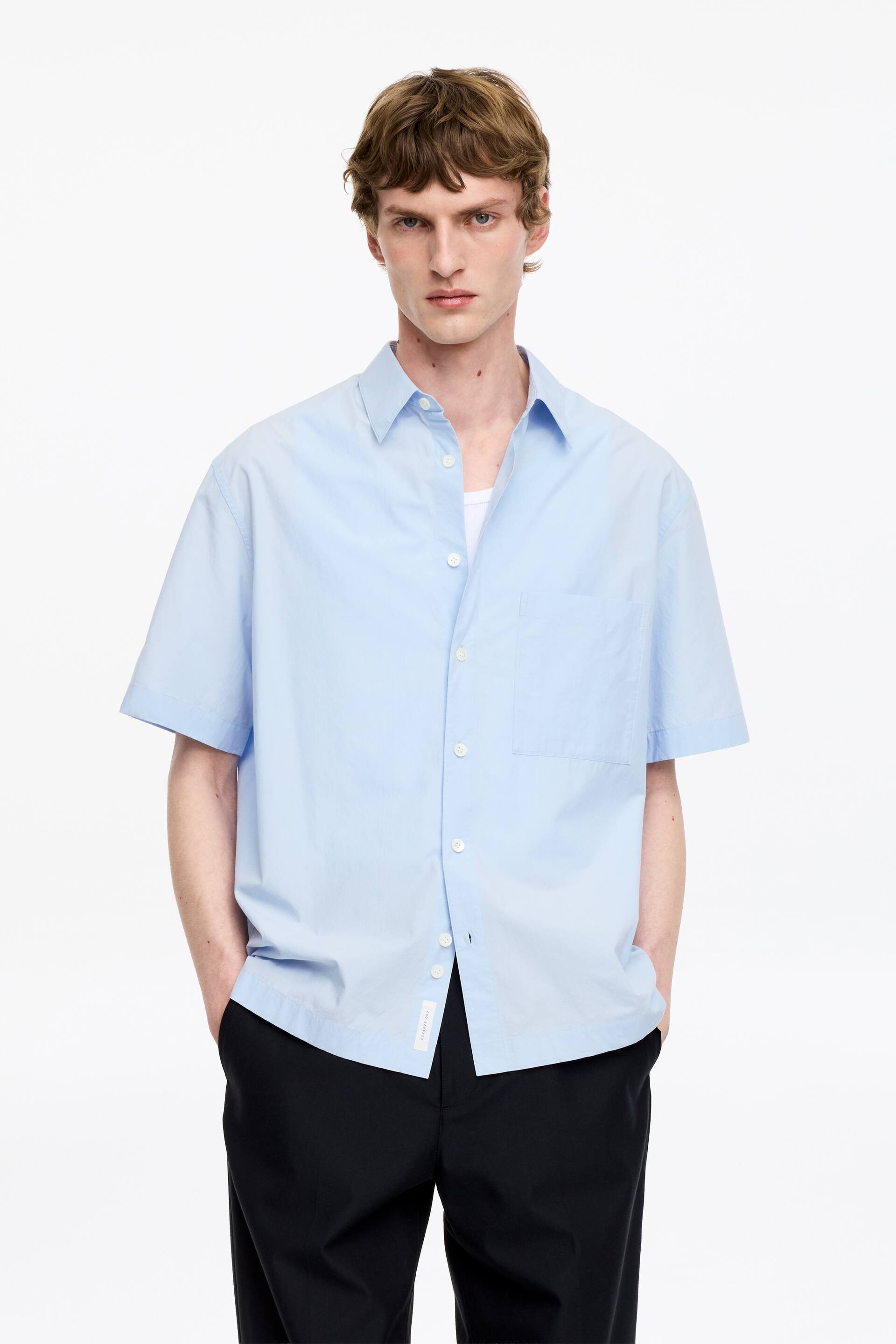 Visualizza immagine più grande: La camicia azzurra a maniche corte presenta una vestibilità oversize, un colletto a punta e una chiusura frontale con bottoni bianchi, mostrata dal davanti. Una tasca a toppa singola è visibile sul petto sinistro e la camicia è stilizzata aperta in alto, rivelando una maglietta bianca sottostante.
