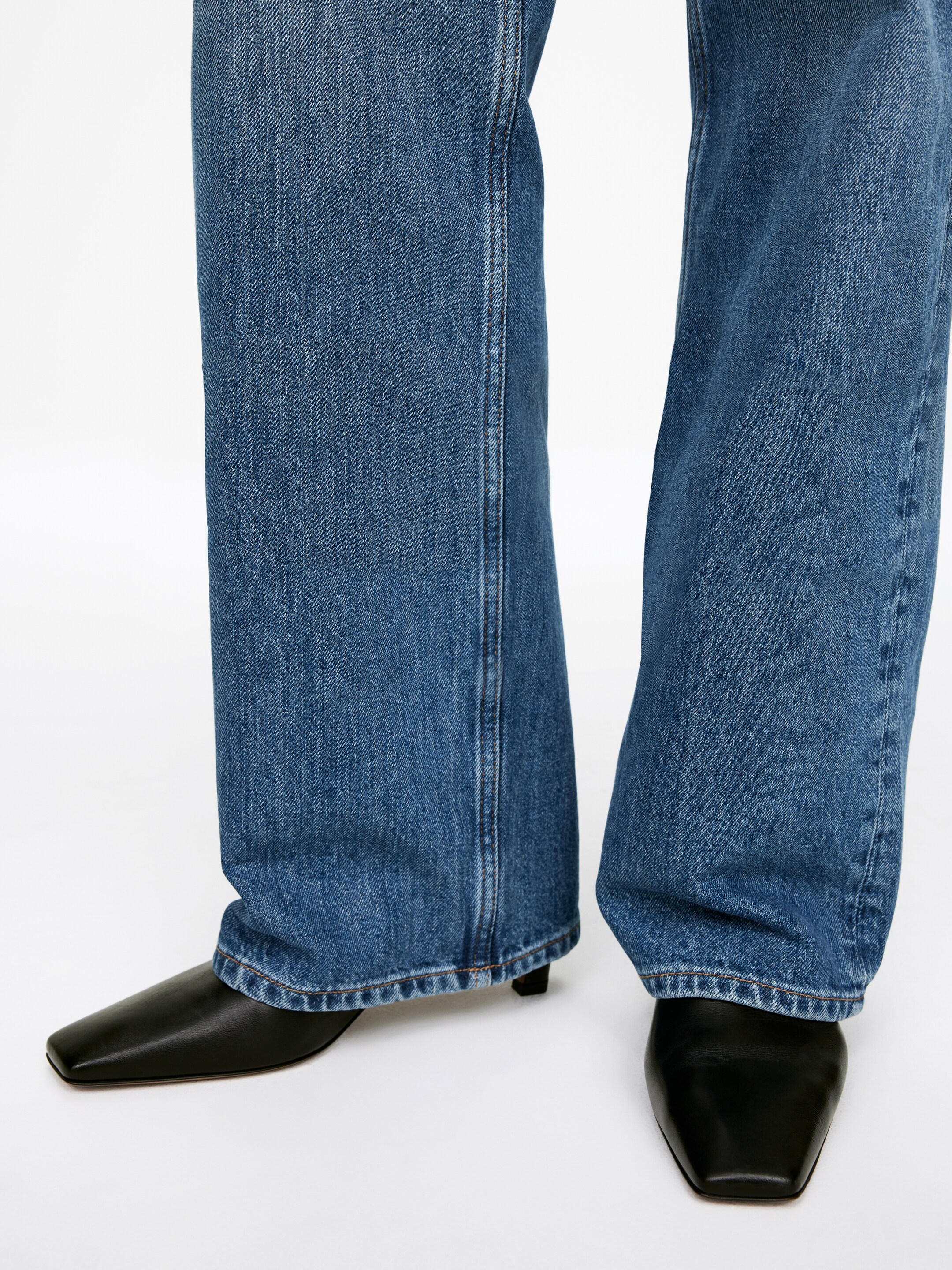 Grotere afbeelding bekijken: Blauwe denim jeans hebben een wijde pasvorm met zichtbare zijnaden en een gestikte zoom, die over zwarte schoenen met vierkante neuzen vallen, welke lage blokhakken hebben.