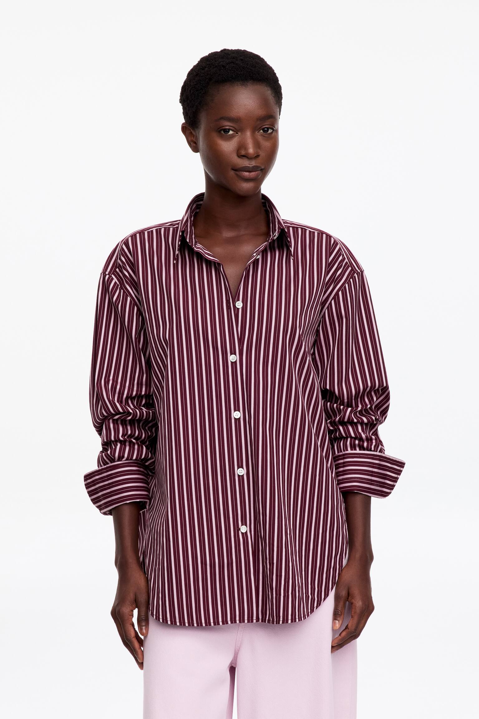Ein langÃ¤rmliges Button-up-Hemd mit burgunder- und weiÃen vertikalen Streifen wird vom Model getragen. Das Hemd hat eine lockere Silhouette mit offenem Kragen, weiÃen KnÃ¶pfen auf der Vorderseite und hochgeschobenen Ãrmeln bis zu den Unterarmen, kombiniert mit hellrosa Hosen. - 1