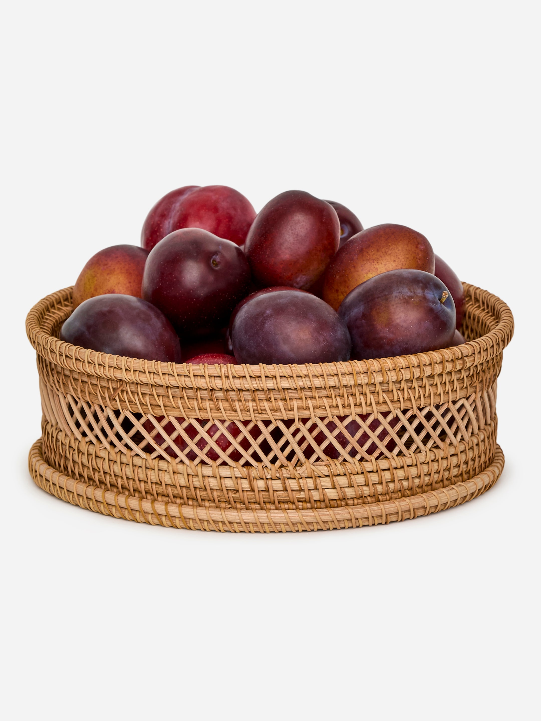 View larger image: A round rattan basket is presented front-on, featuring a solid woven base and top rim, with an open diamond lattice weave forming its sides. It is holding a pile of dark red and purple plums.