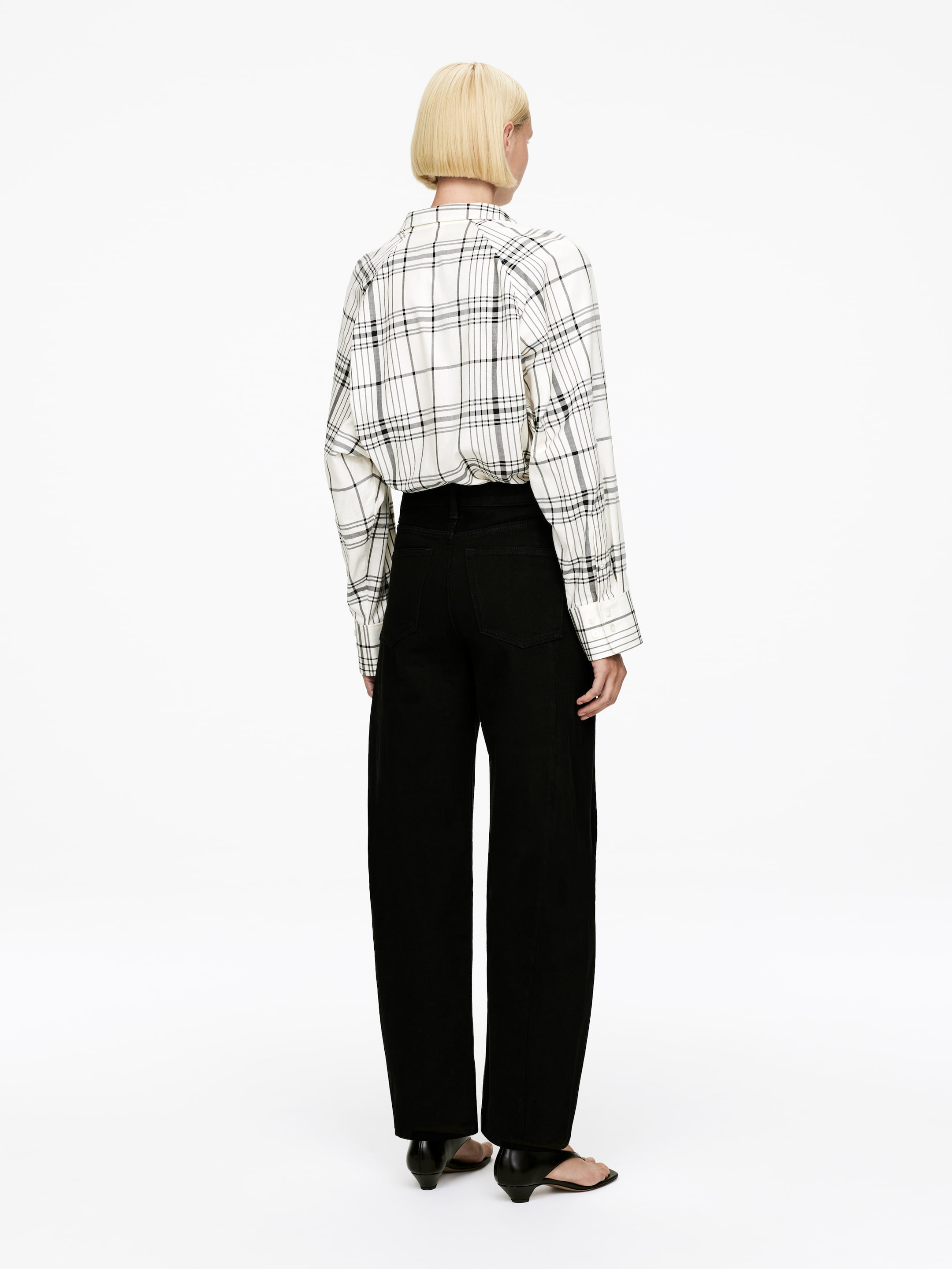 Visualizza immagine più grande: Una camicia a quadri bianca, nera e grigia con bottoni è indossata, infilata in pantaloni neri a gamba larga. I pantaloni presentano dettagli di cuciture visibili sul retro delle gambe e sono abbinati a mule nere aperte in punta, il tutto visto da dietro.