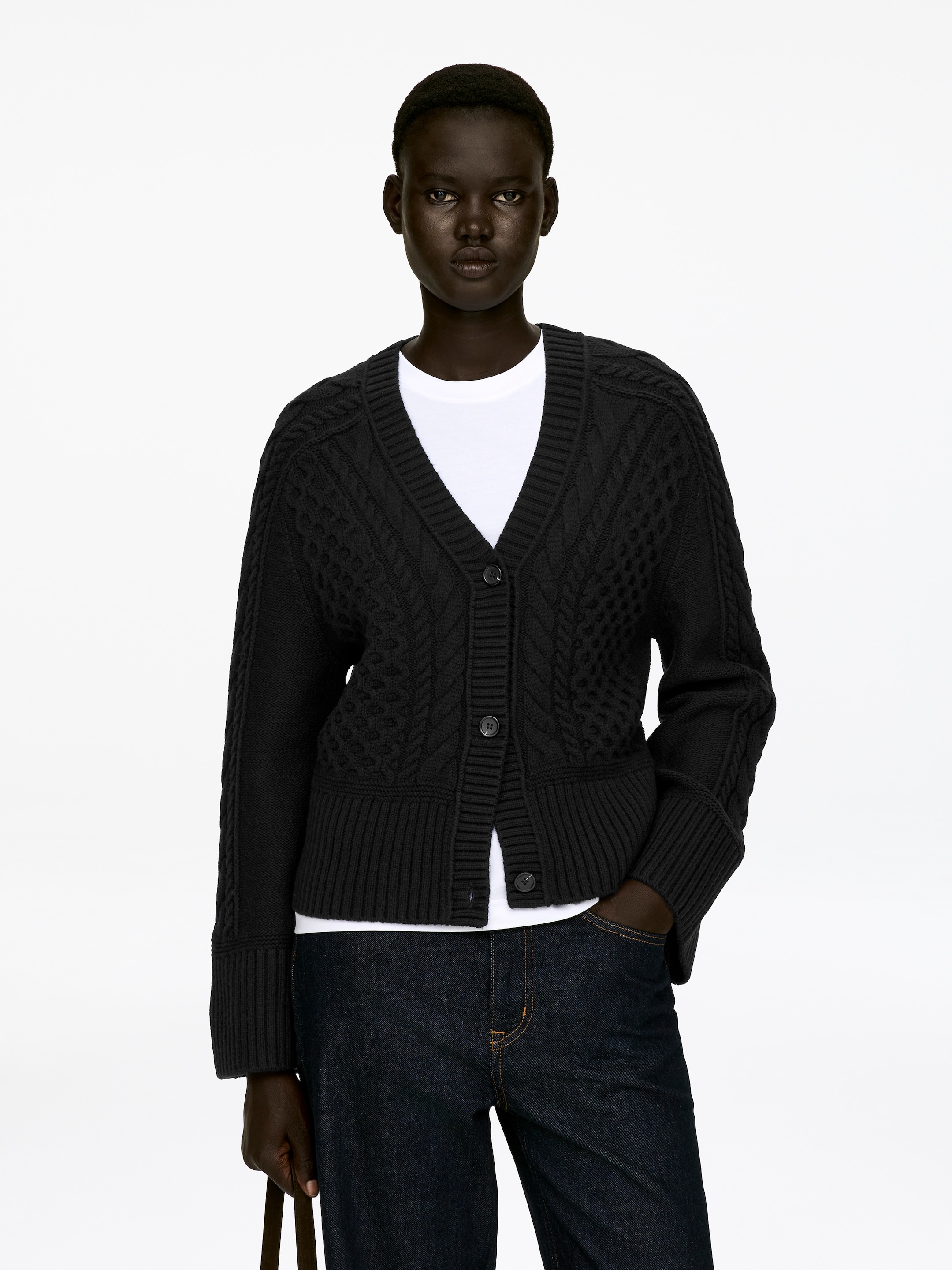 Grösseres Bild ansehen: Eine schwarze Zopfstrickjacke mit V-Ausschnitt, Dreiknopfleiste und gerippten Abschlüssen wird vom Model über einem weißen T-Shirt getragen. Das Model ruht eine Hand in einer Tasche dunkler Jeans.