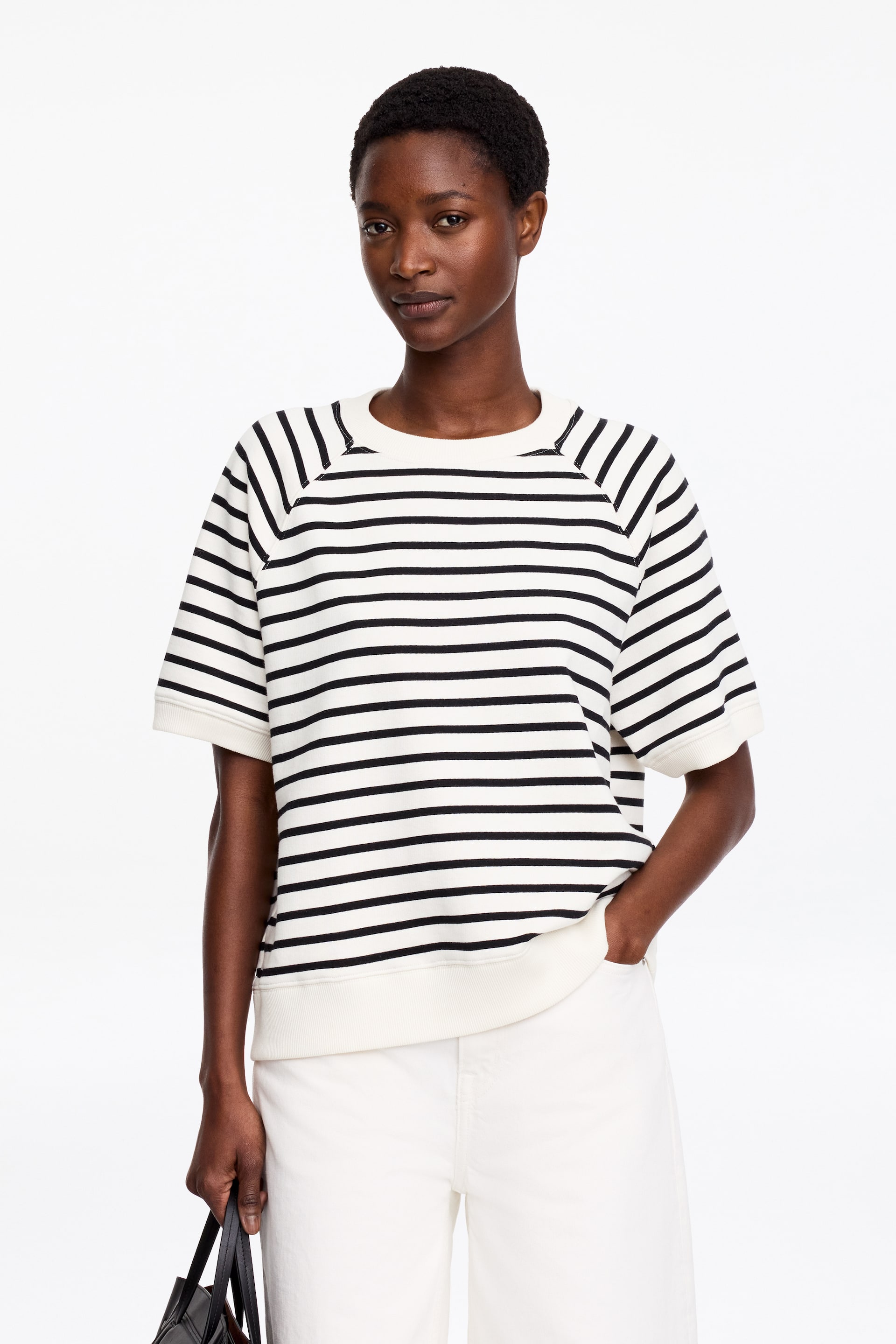 Agrandir l'image: Un sweat à manches courtes, avec des rayures horizontales noires et crème, un col rond, des manches raglan et des finitions en maille côtelée aux poignets et à l'ourlet, est porté par le mannequin. Le mannequin le porte avec un pantalon blanc, une main dans la poche du pantalon et l'autre tenant un sac noir.