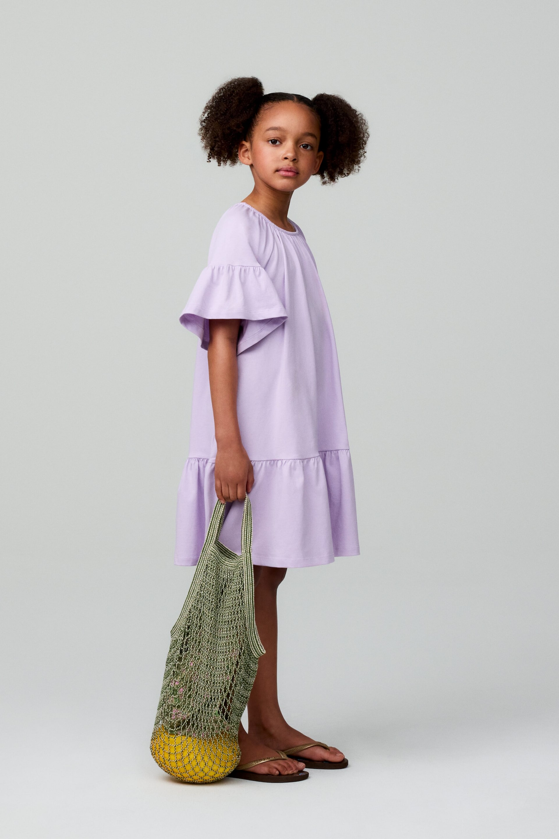 Größeres Bild ansehen: Das hellviolette Kleid zeichnet sich durch einen gerafften Rundhalsausschnitt und kurze Rüschenärmel aus, die in einen gestuften Rock mit Rüschenabschluss übergehen.